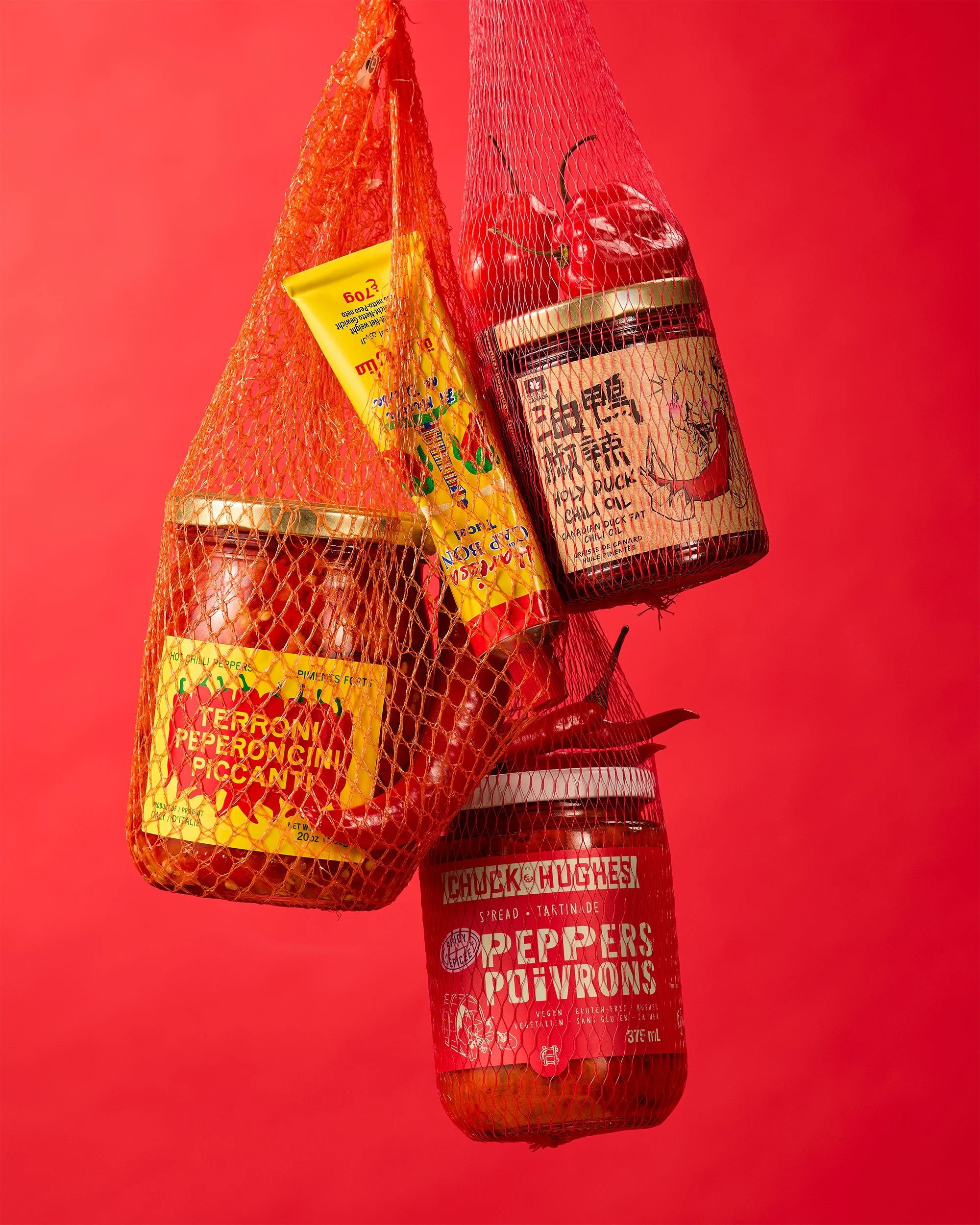 Still life creative photography prop styled and art directed by rayna marlee. A red mesh bag hanging against a red background, containing jars labeled 'Peppers Powders,' a can labeled 'Holy Duck Chili Oil,' and a package of peppers.
