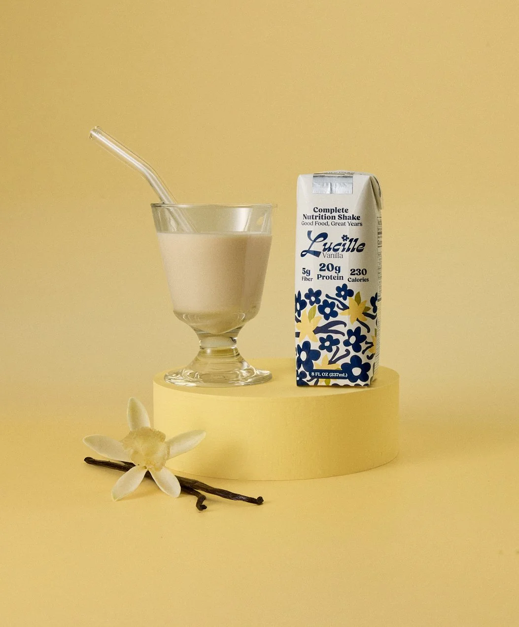 Still life creative photography prop styled by rayna marlee. A glass of vanilla protein shake with a straw, a carton of Lucille vanilla protein beverage, a vanilla flower, and a vanilla bean on a small yellow platform against a yellow background.