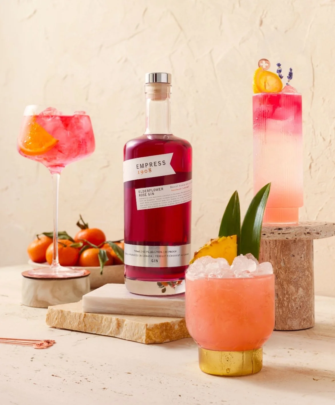 Drink creative photography prop styled by rayna marlee.A  bottle of Empress Elderflower Rose Gin surrounded by three colorful cocktails garnished with fruit, set against a textured beige background.