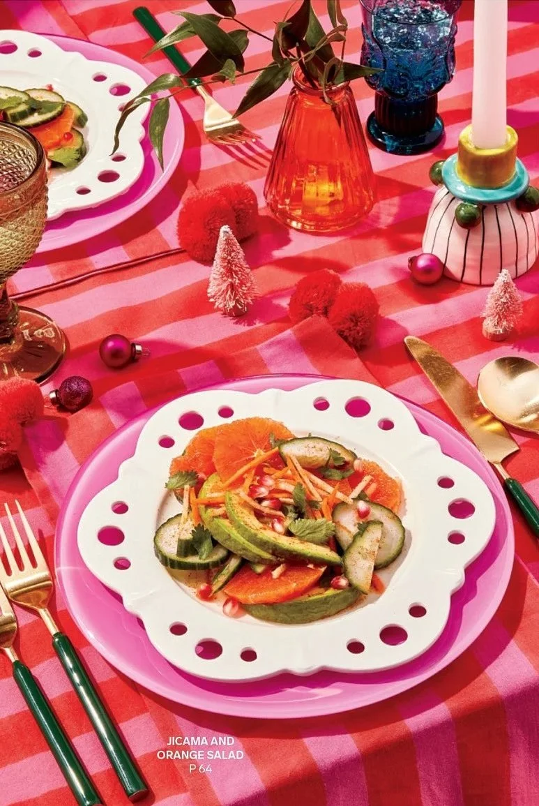 A colorful table setting with a plate of cucumber and orange salad garnished with herbs and pomegranate seeds, surrounded by festive decorations like small pink trees, Christmas ornaments, candles, and vibrant glassware, on a pink and red striped tab