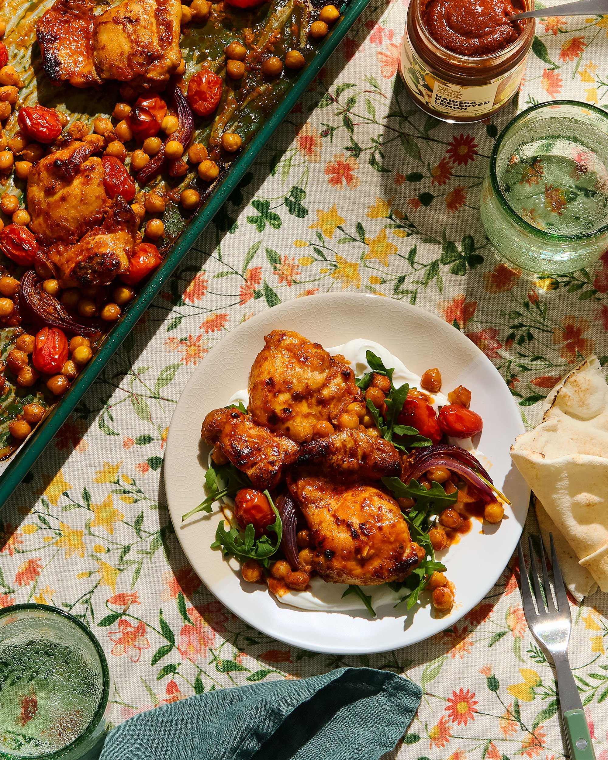 Food creative photography prop styled and art directed by rayna marlee. Plate of grilled chicken with chickpeas, cherry tomatoes, and arugula salad on a floral tablecloth, with a jar of red sauce, two glasses of sparkling water, and naan bread