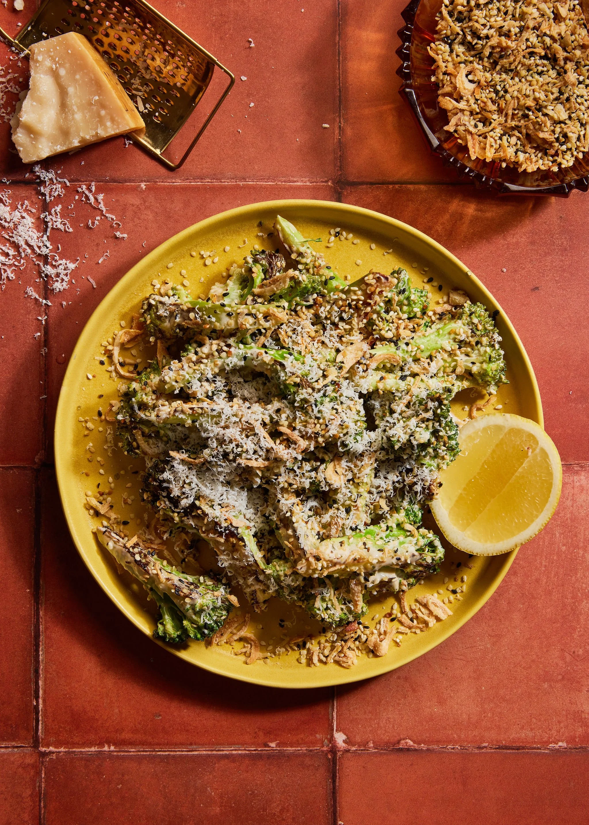 Food creative photography prop styled and art directed by rayna marlee. A yellow plate of sautéed vegetables topped with shredded cheese and sesame seeds, garnished with lemon wedges on a red tiled surface, with grated cheese