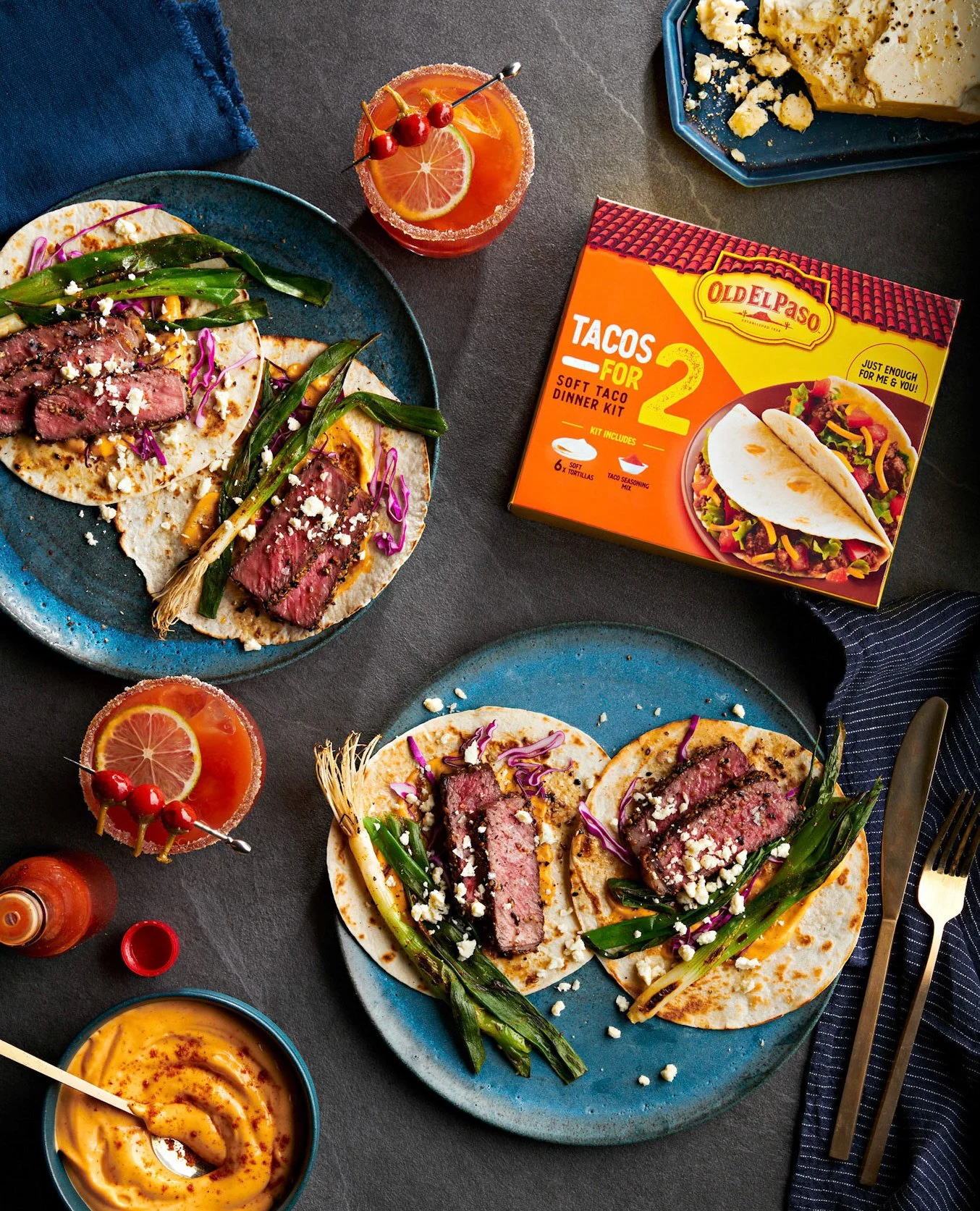 Two plates with beef tacos topped with green onions and crumbled cheese, two cocktails with lemon slices and cherries, a bowl of sauce, a box of taco dinner kit, and utensils on a dark table.