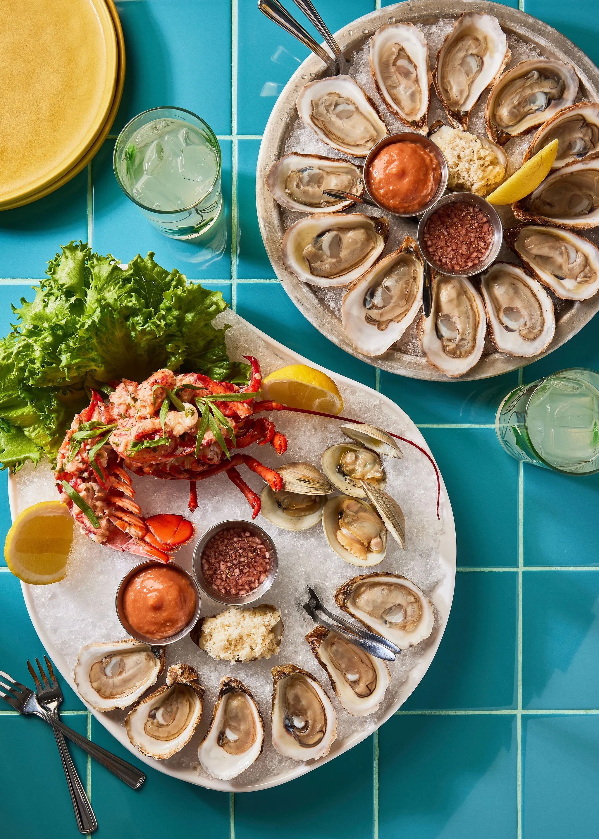 HEYDAYS RESTAURANT COOKBOOK at The June Motel. A styled photographed top view of two plates of oysters on ice, with lemon wedges, cocktail sauce, and horseradish, placed on a bright blue tiled table, with glasses of water nearby.