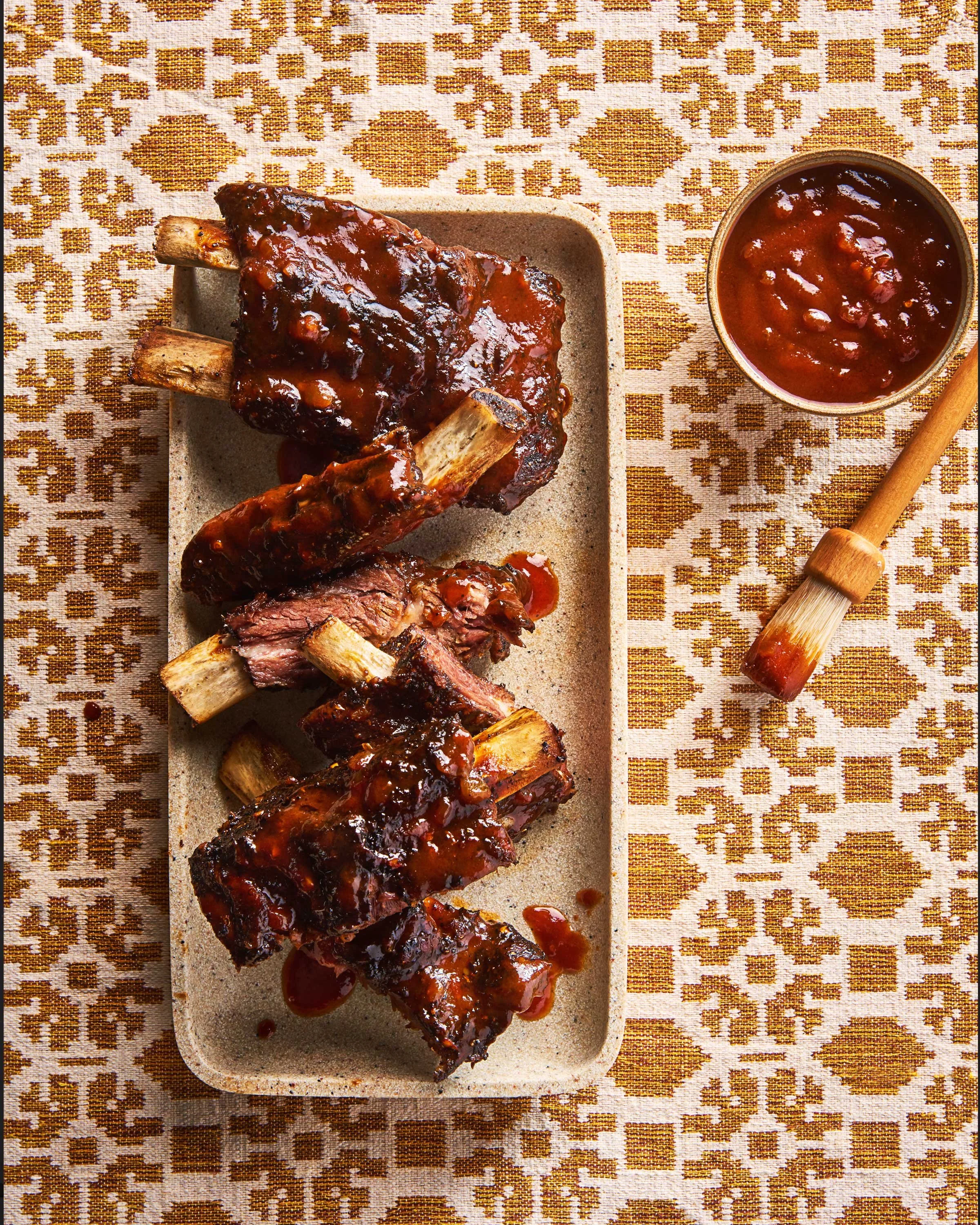 Cover of a cookbook titled "Eat, Habibi, Eat!" by Shahir Massoud. Art direction. BBQ pork ribs with sauce on a rectangular beige plate, accompanied by a small bowl of barbecue sauce, on a patterned tablecloth with a wooden basting brush nearby.