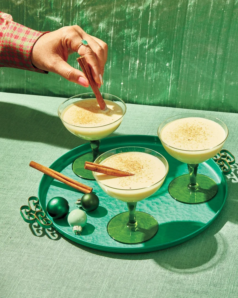 Three glasses of eggnog garnished with cinnamon sticks on a green tray, with holiday ornaments on a green table, against a green backdrop.
