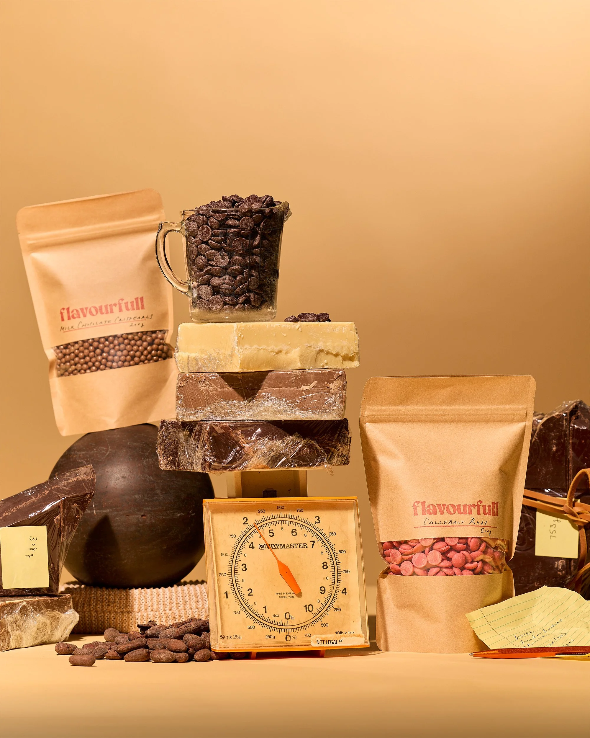 Still life creative photography prop styled and art directed by rayna marlee. Display of various chocolate ingredients including cocoa beans, cocoa butter, chocolate bars, and packaged chocolate products on a kitchen scale