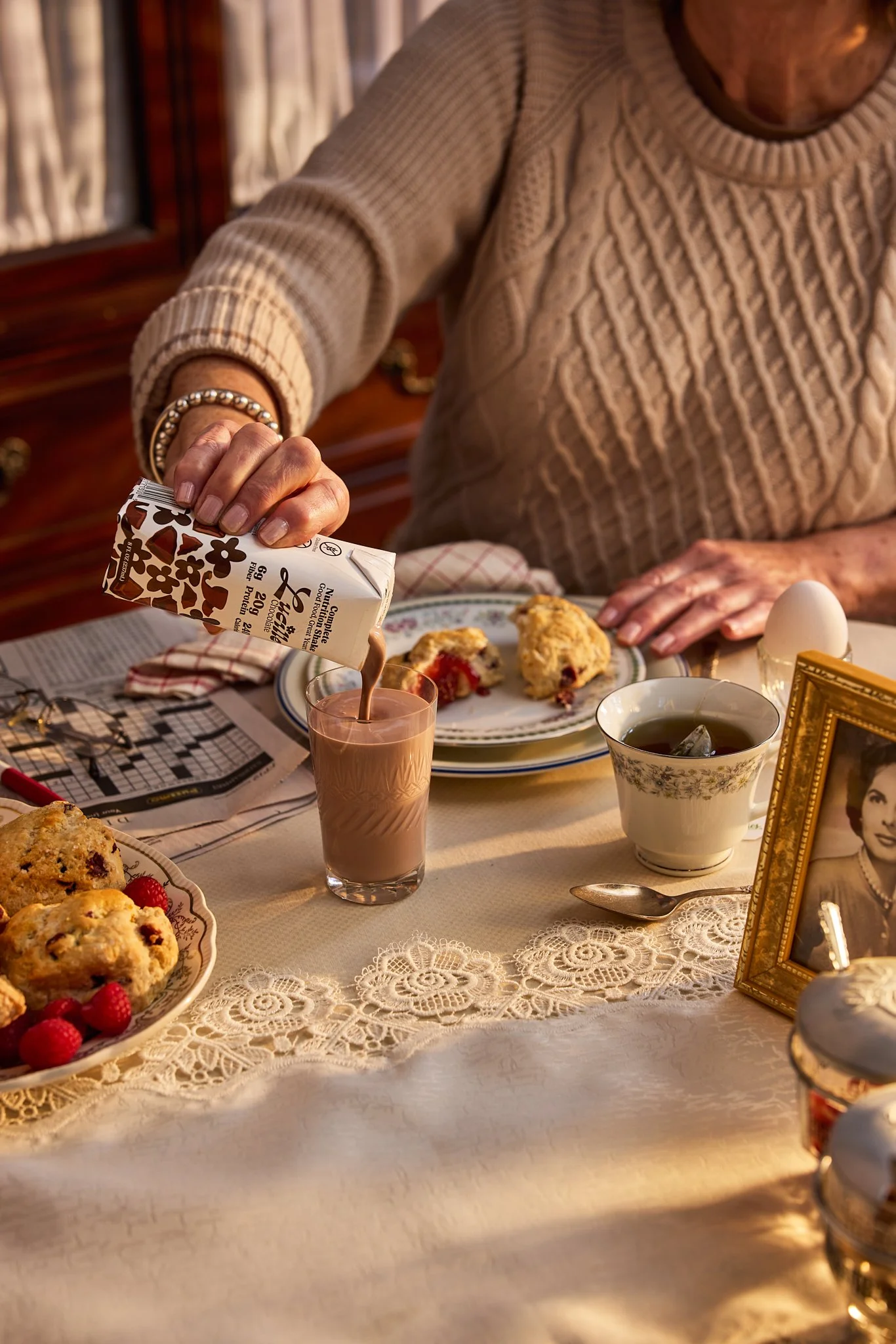 Prop styling by rayna marlee. Photography of An elderly woman pouring chocolate milk into a glass at a table set for breakfast with scones, raspberries, tea, an egg, a steaming cup, a framed photo, and a lace tablecloth.