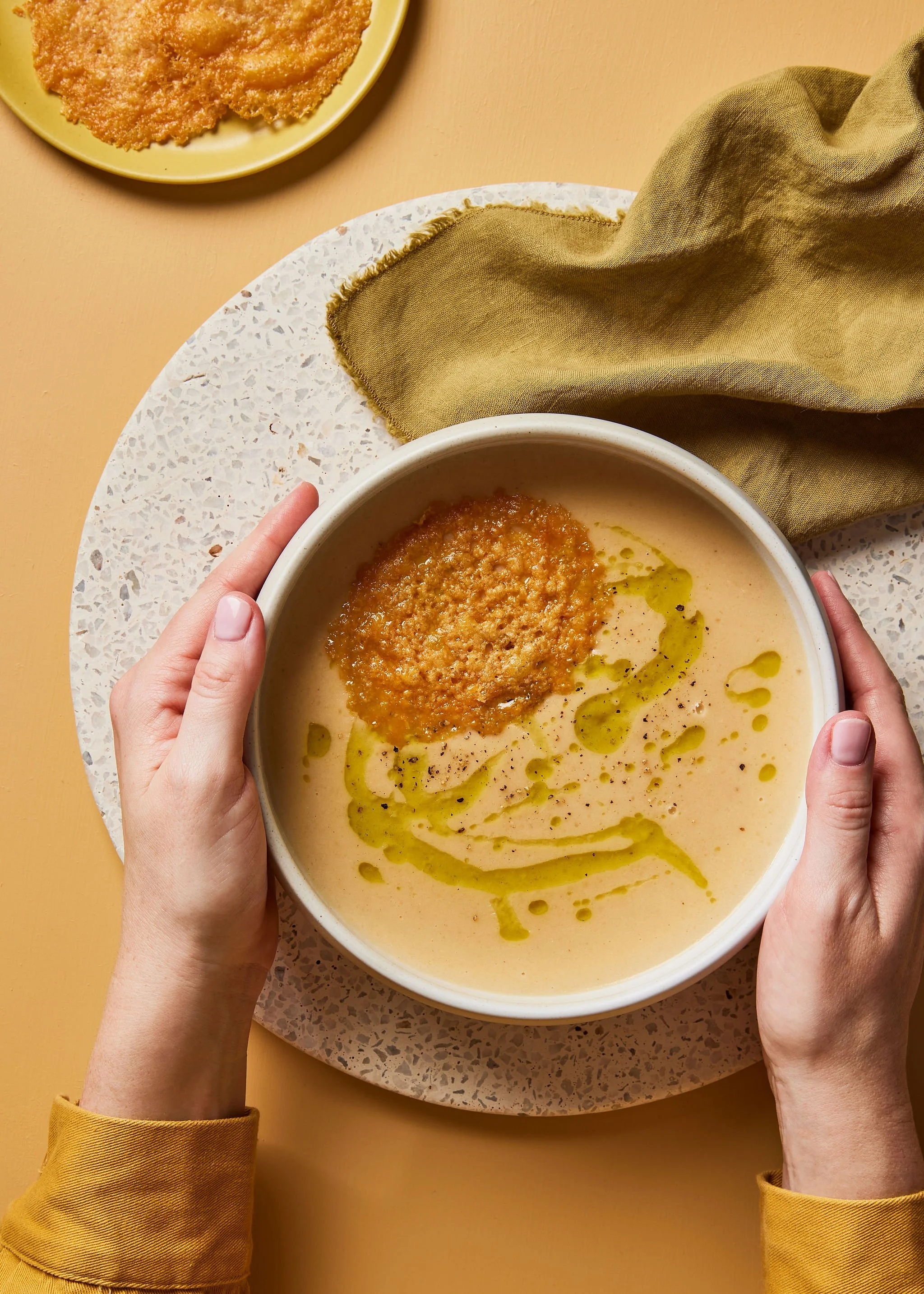 Food creative photography prop styled and art directed by rayna marlee for Hey Days cookbook. A bowl of creamy soup garnished with a toasted bread slice, drizzled with olive oil and black pepper, held by two hands.
