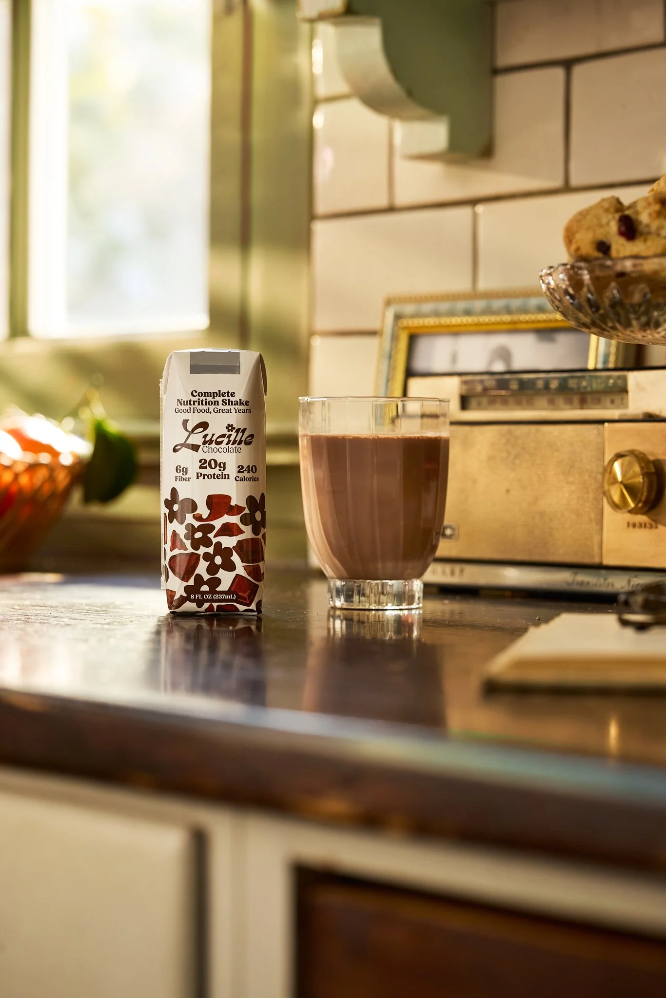 A glass of chocolate protein shake on a kitchen counter next to a carton of Lucille chocolate nutrient shake, with a toaster oven and a framed photo in the background.