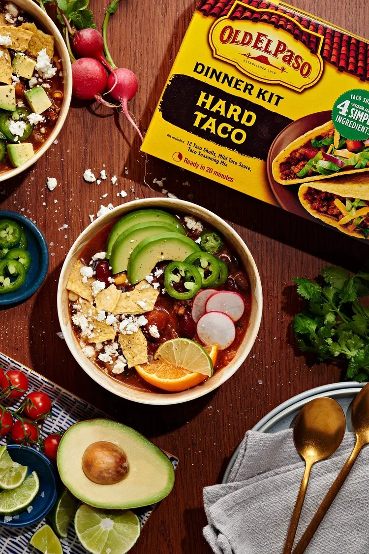 A bowl of Mexican-style chili with sliced avocado, radishes, lime, and tortilla chips. Surrounding bowls contain sliced jalapeños, cherry tomatoes, lime wedges, and radishes. A box of Old El Paso dinner kit for hard tacos is on the table.