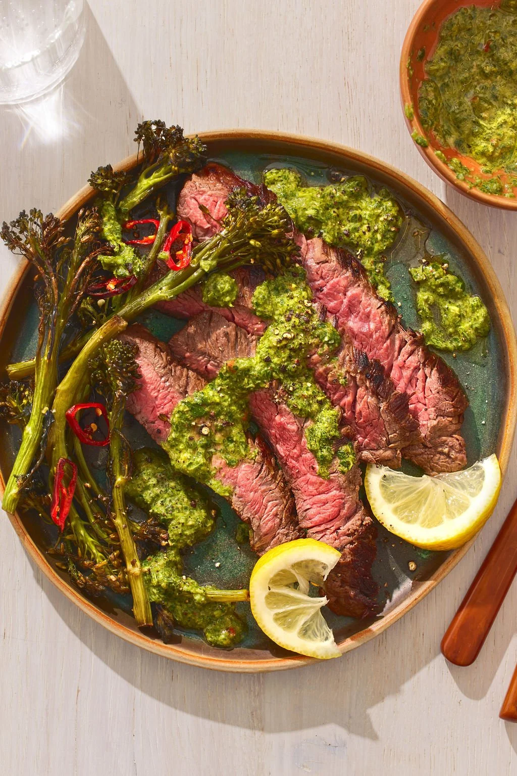Cover of a cookbook titled "The Young Forever Cookbook" by Mark Hyman, MD, art directed and prop styled by rayna marlee. Slices of cooked steak topped with green chimichurri sauce, served with grilled broccolini and lemon wedges on a ceramic plate.