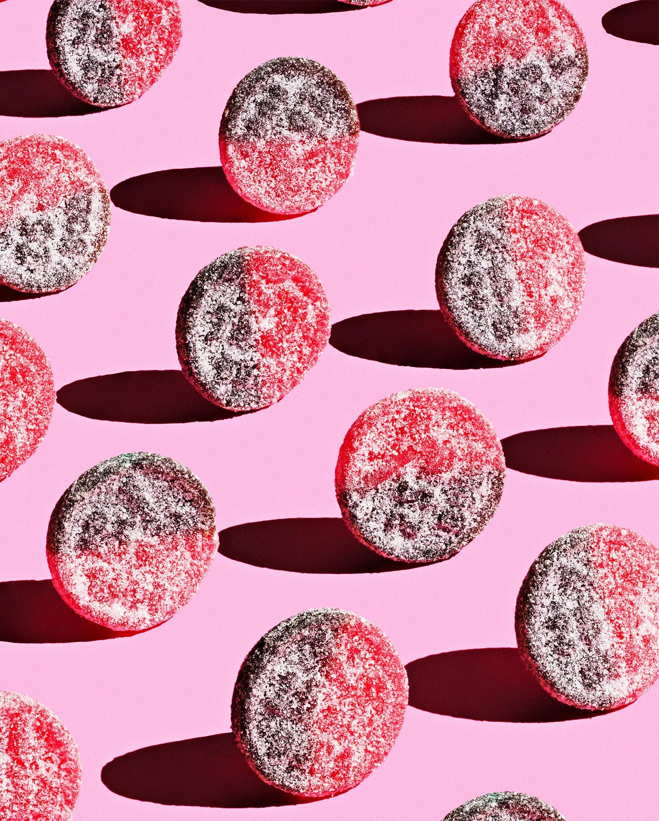 Candy creative photography prop styled and art directed by rayna marlee for Sukker Baby. Red and black round candies covered in sugar on a pink background. Shadows cast by the candies.