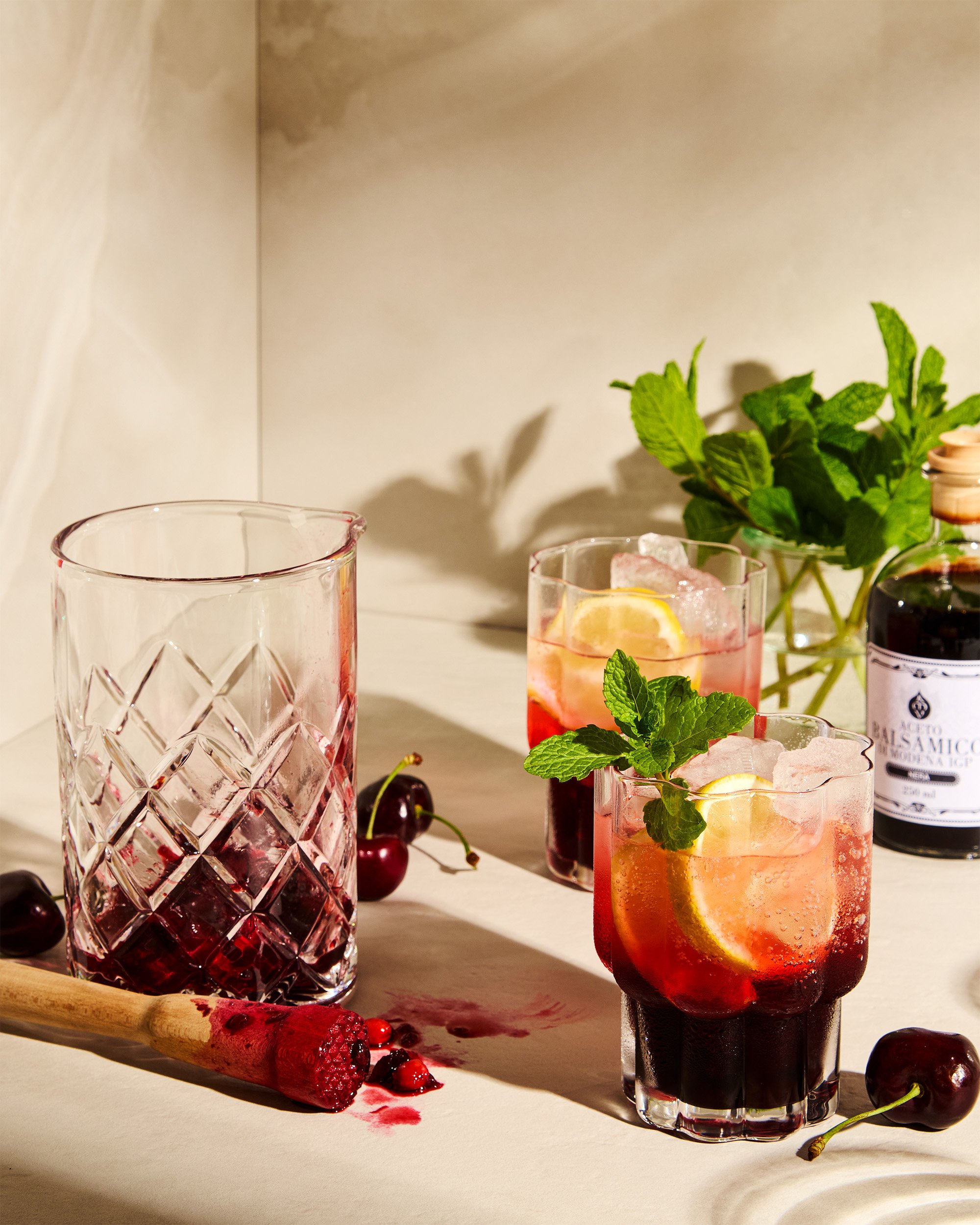 drink photography prop styled and art directed by rayna marlee Two glasses of cherry and lemon cocktails with mint garnishes, a cutting board with a cherry and juice spill, a bunch of cherries, a jar of balsamic vinegar, and a plant of fresh mint..