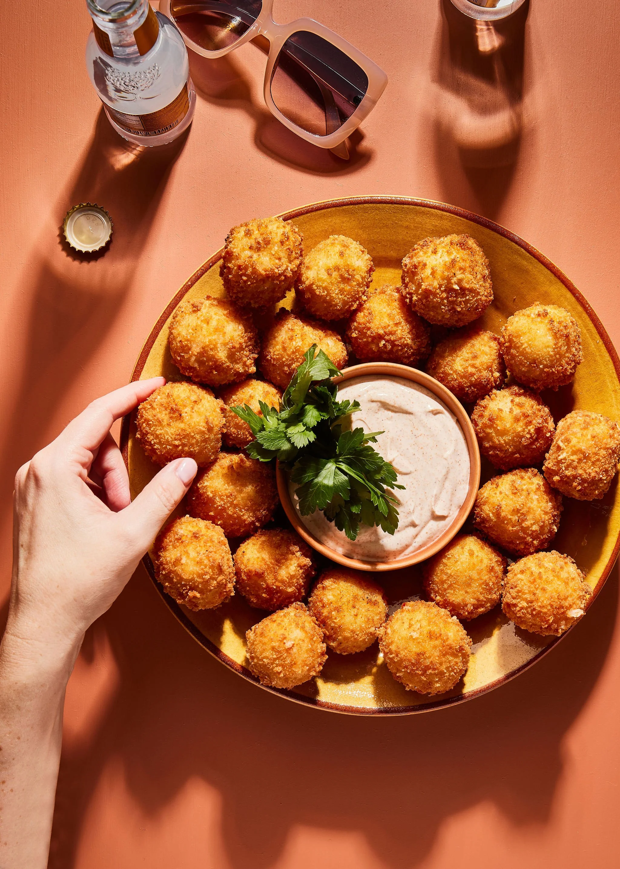 Food photography. A hand reaches for a plate of round, breaded, fried appetizers arranged around a bowl of dipping sauce garnished with parsley, on a pink surface, with sunglasses, a water bottle, and a glass bottle in the background.