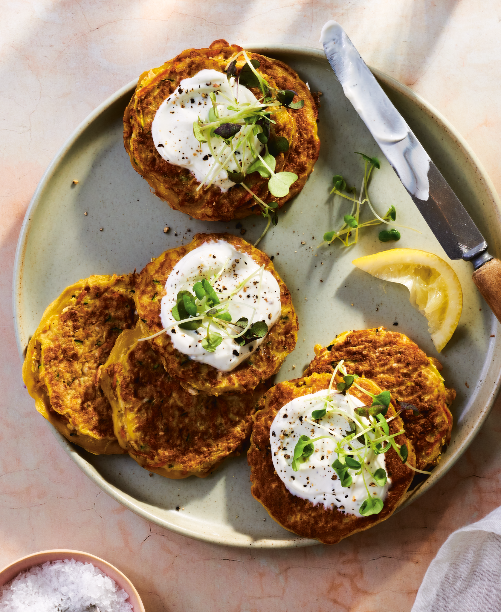 Photography cover of a cookbook titled 'Good Food, Good Mood'  with a late of four veggie patties topped with sour cream and microgreens, lemon wedge, and a knife on a light-colored surface. Prop styled by rayna marlee.