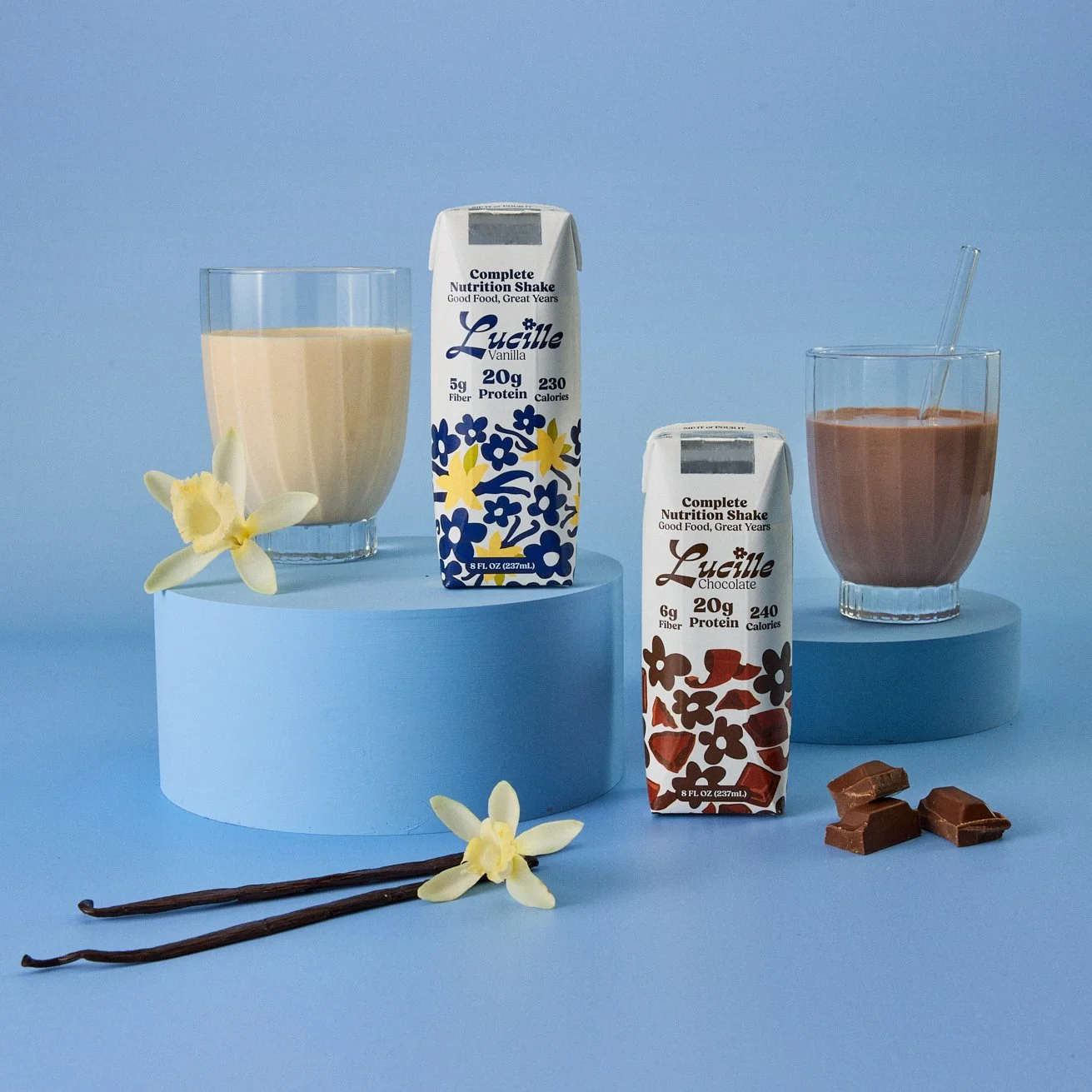 photography prop styled by rayna marlee Two glasses of flavored milk, one vanilla and one chocolate, with cartons of each flavor in the middle, surrounded by vanilla flowers, chocolate pieces, and two vanilla beans against a blue background.