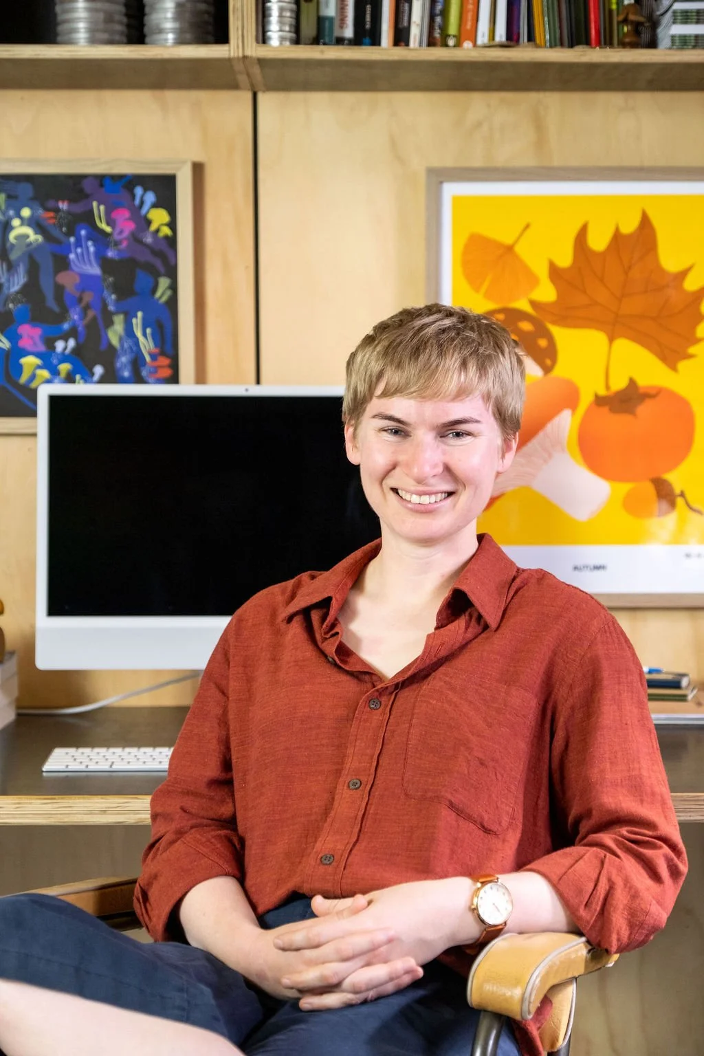 A smiling blonde-haired woman sits in front of her desktop computer and workspace.