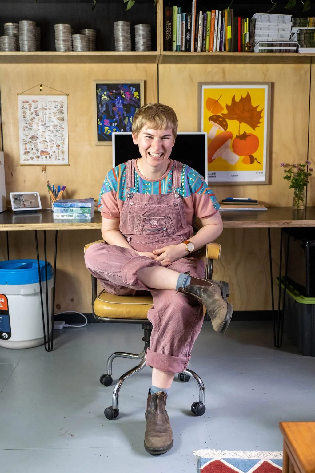 A smiling blonde-haired woman in pink corduroy overalls sits at her desk.