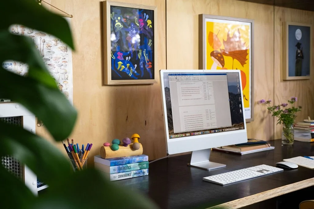 A beautiful desk space with a desktop computer, framed art prints of fungi, a stack of books, and a potted plant