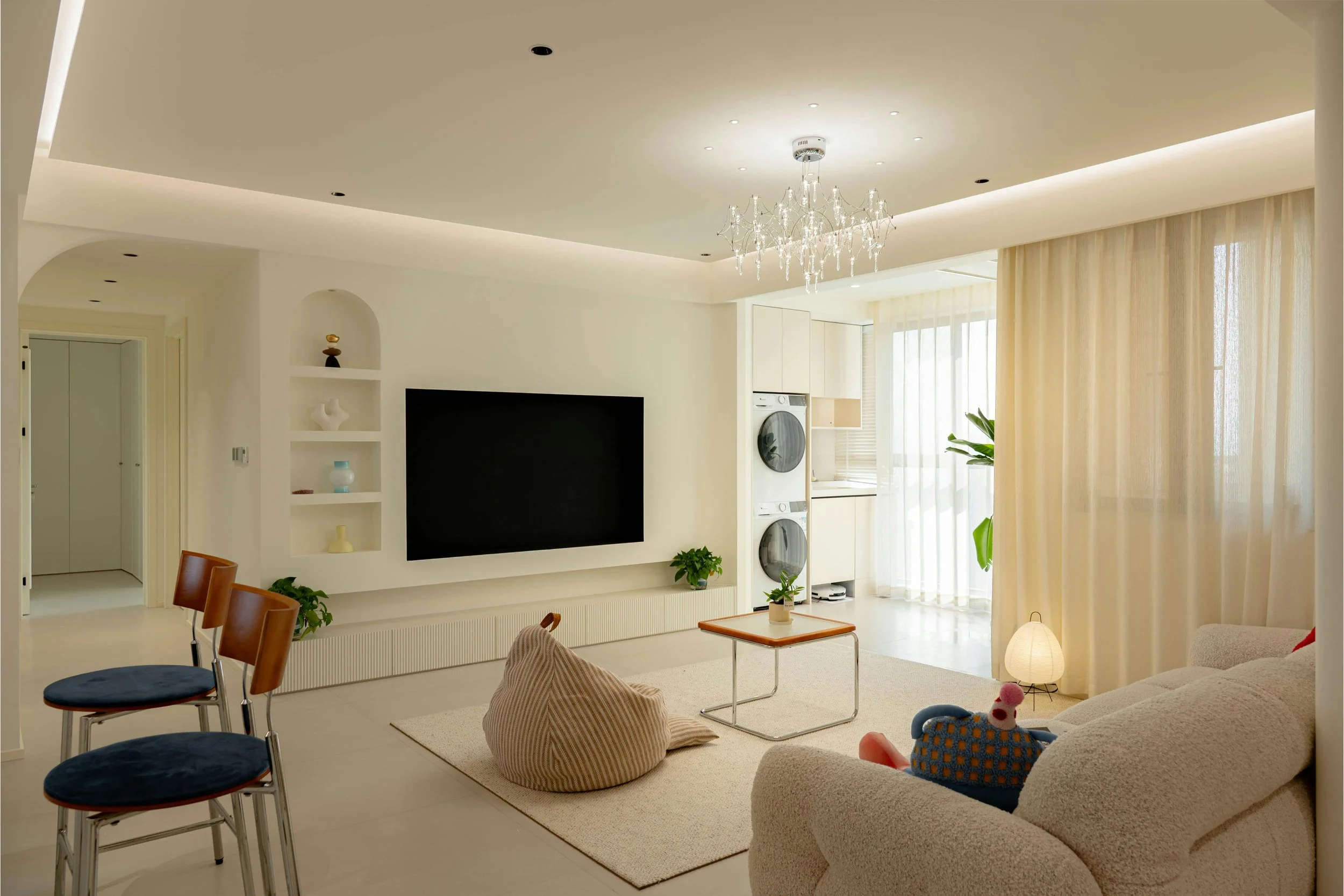 Modernes Wohnzimmer mit weißer Couch, Teppich, kleinen Tisch, TV, Stehlampe, Wasserkocher, Bügeleisen, Fenster mit Vorhängen, dekorative Pflanzen und bunte Kissen.