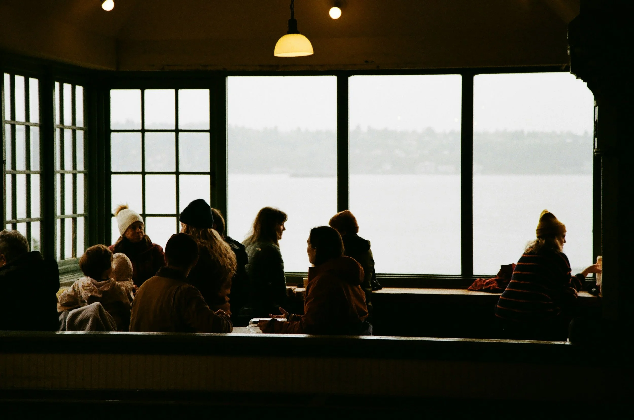 Several people sitting and standing near large windows with a lakeside view, some wearing winter hats, inside a dimly lit cafe or restaurant.