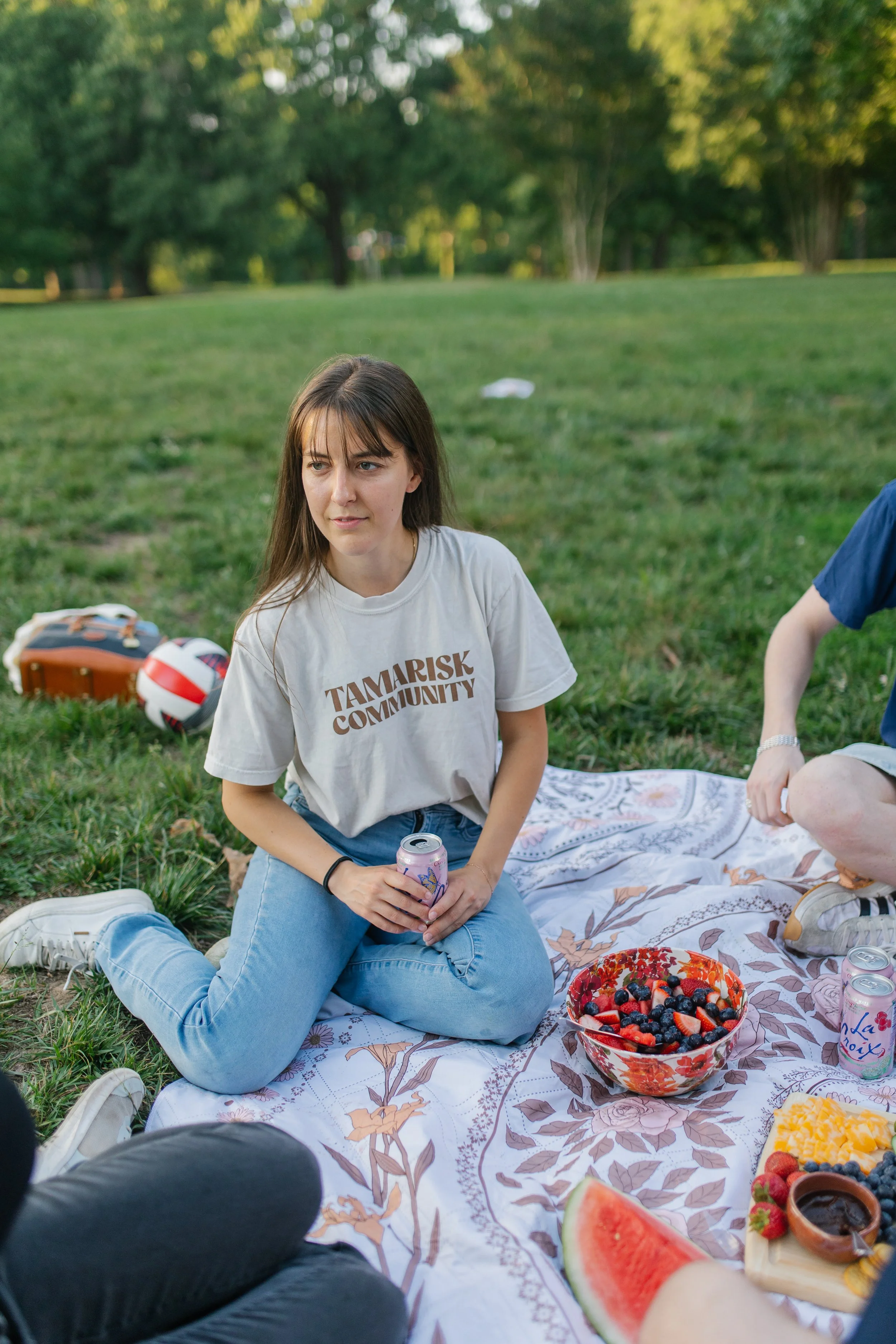 A young woman sitting on a picnic blanket in a park, holding a soda can, with a bowl of mixed berries nearby, during a sunny day.