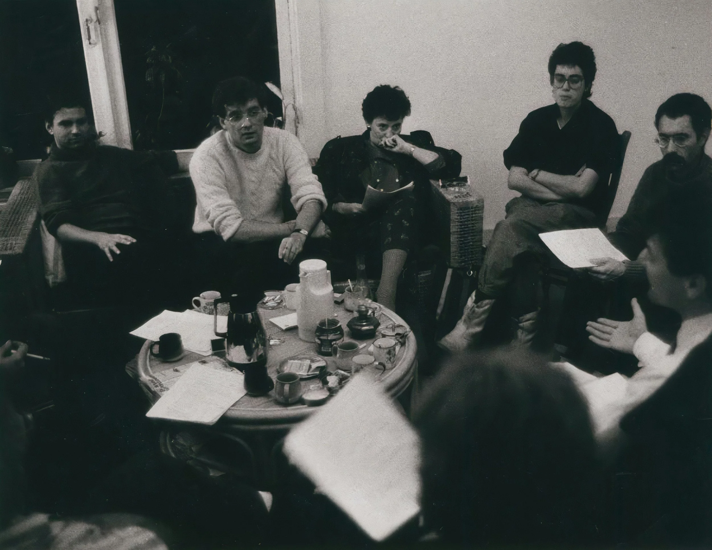 Black-and-white photo of six people sitting around a table with food and drinks, with a window in the background.