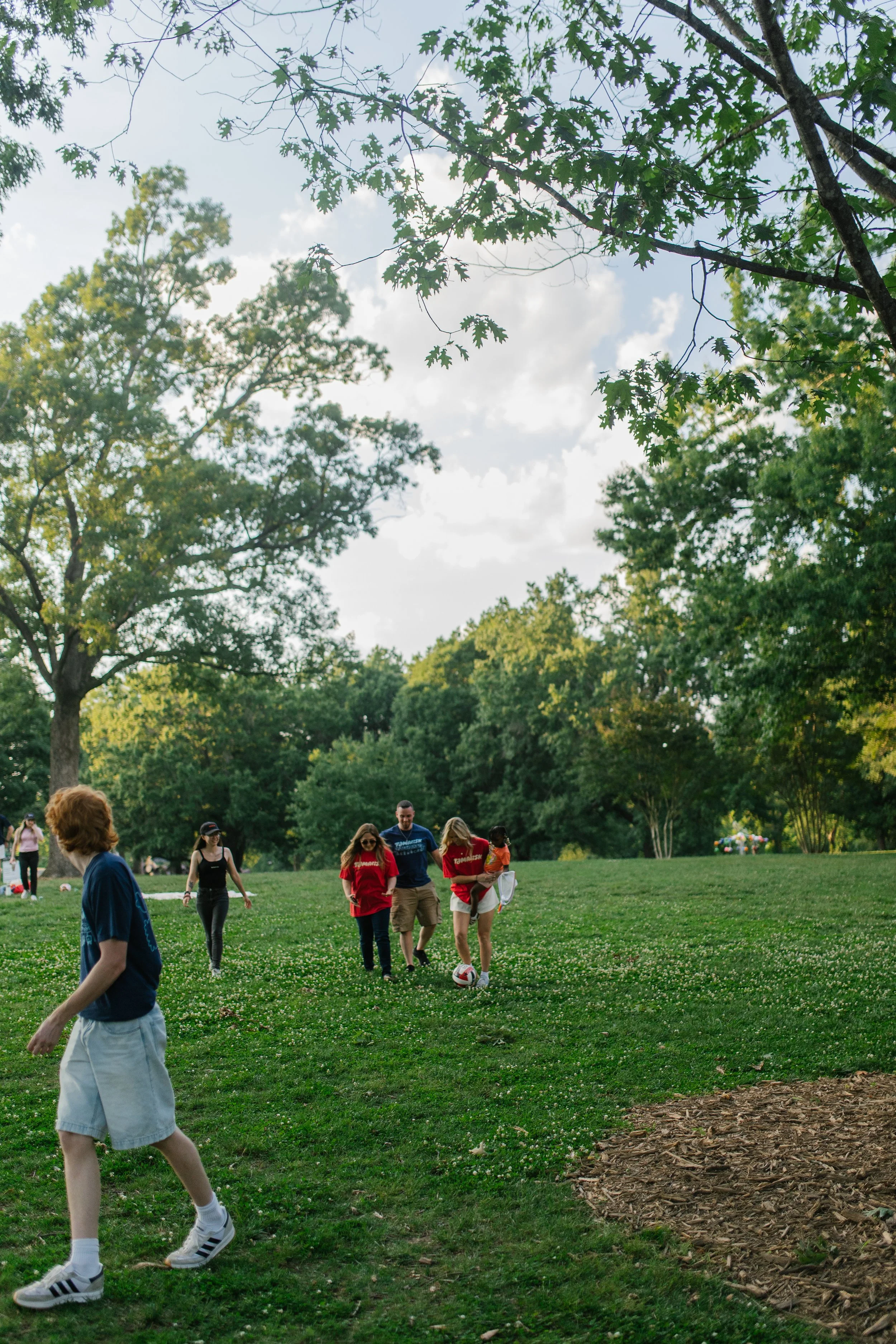 People playing soccer in a park with large trees and a grassy field.
