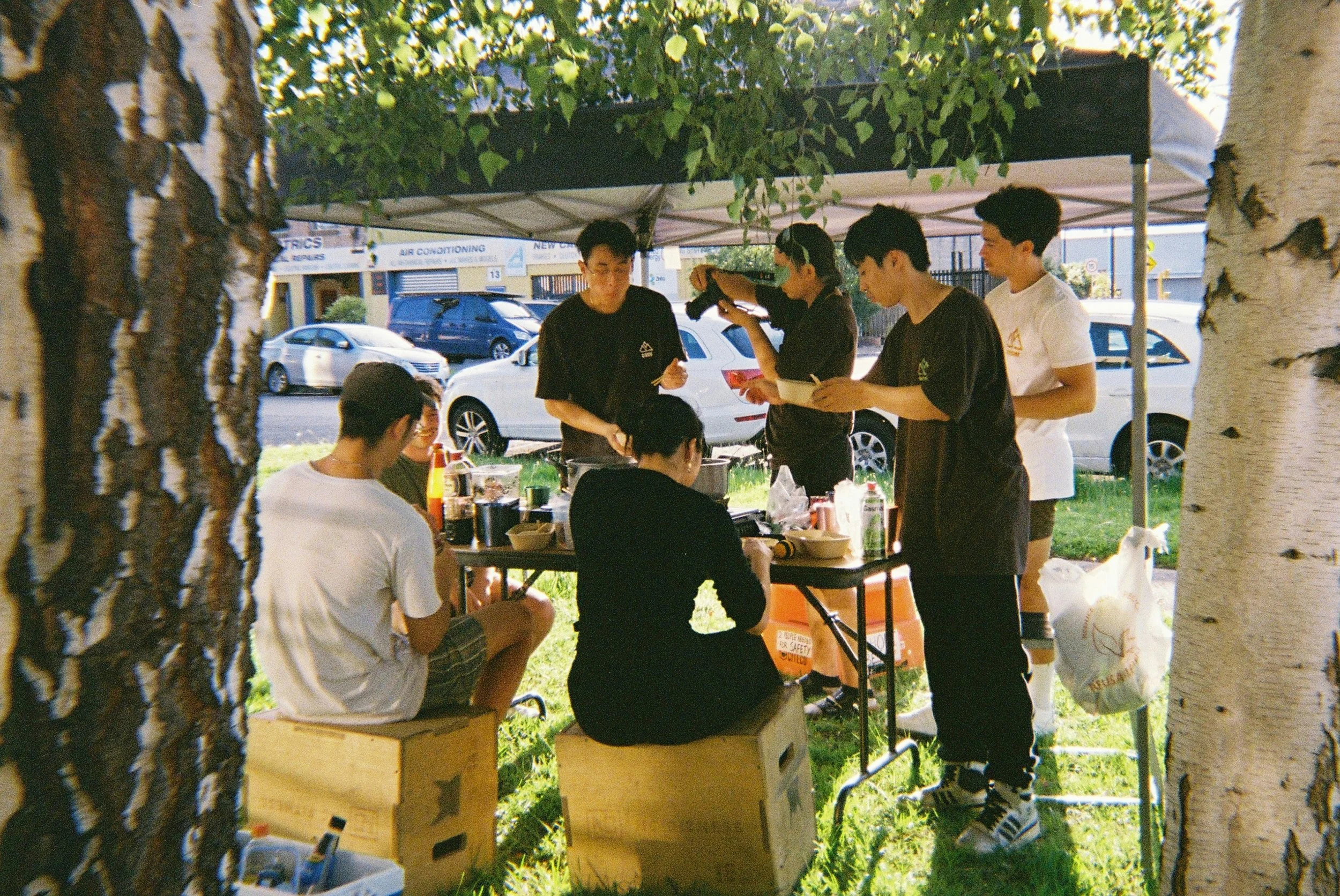 Group of people preparing food at an outdoor stall under a canopy, with cars and a building in the background, framed by tree trunks.