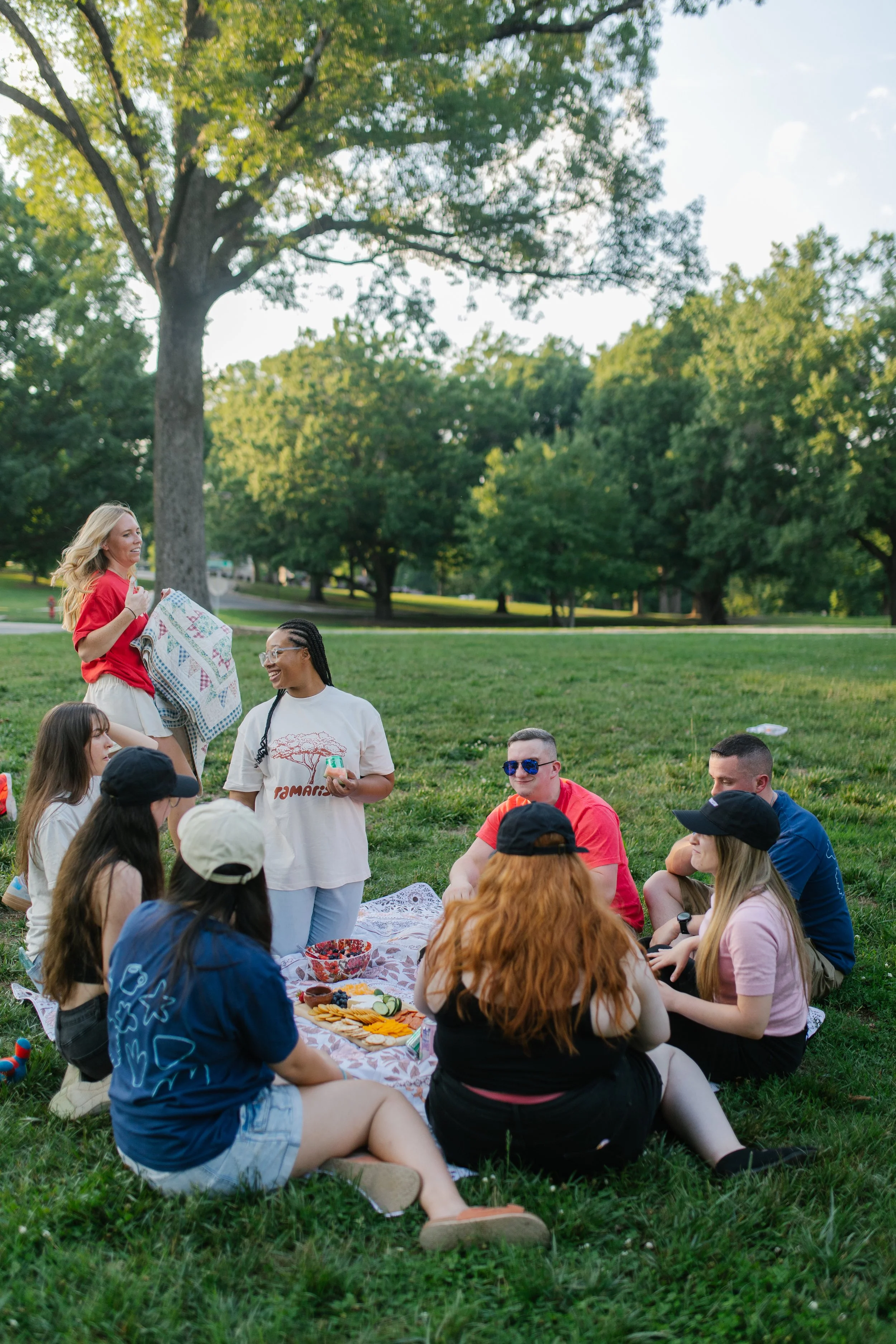 A group of people, mostly young women, are sitting on a blanket having a picnic in a park. They are surrounded by trees and green grass. Some are wearing hats and sunglasses. Food and snacks are spread out on the blanket. A woman in a red shirt is standing and talking, holding a quilt or blanket.