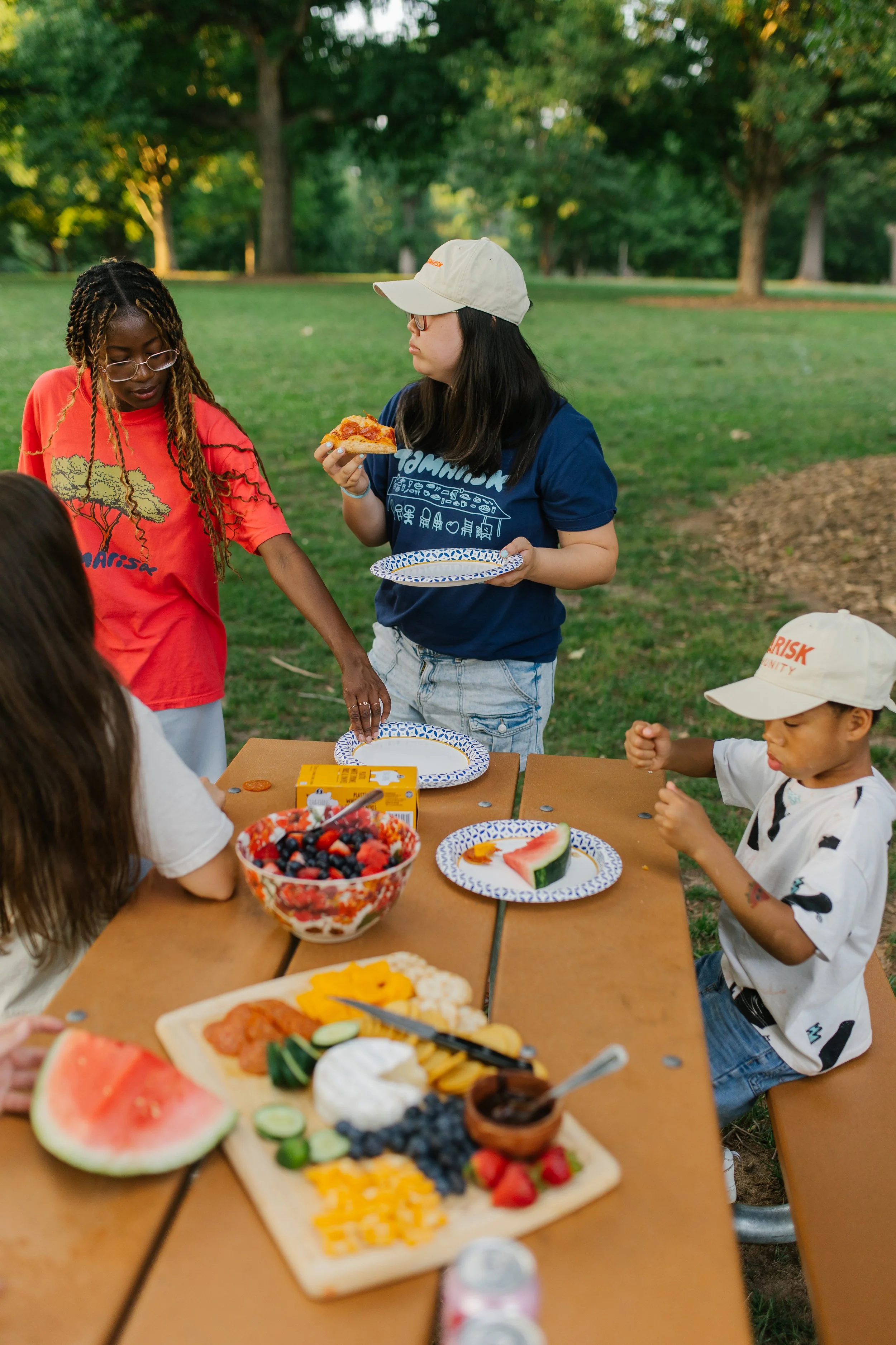 People enjoying a picnic in a park with various foods, including fruit, pizza, and desserts, on a wooden table, with trees in the background.