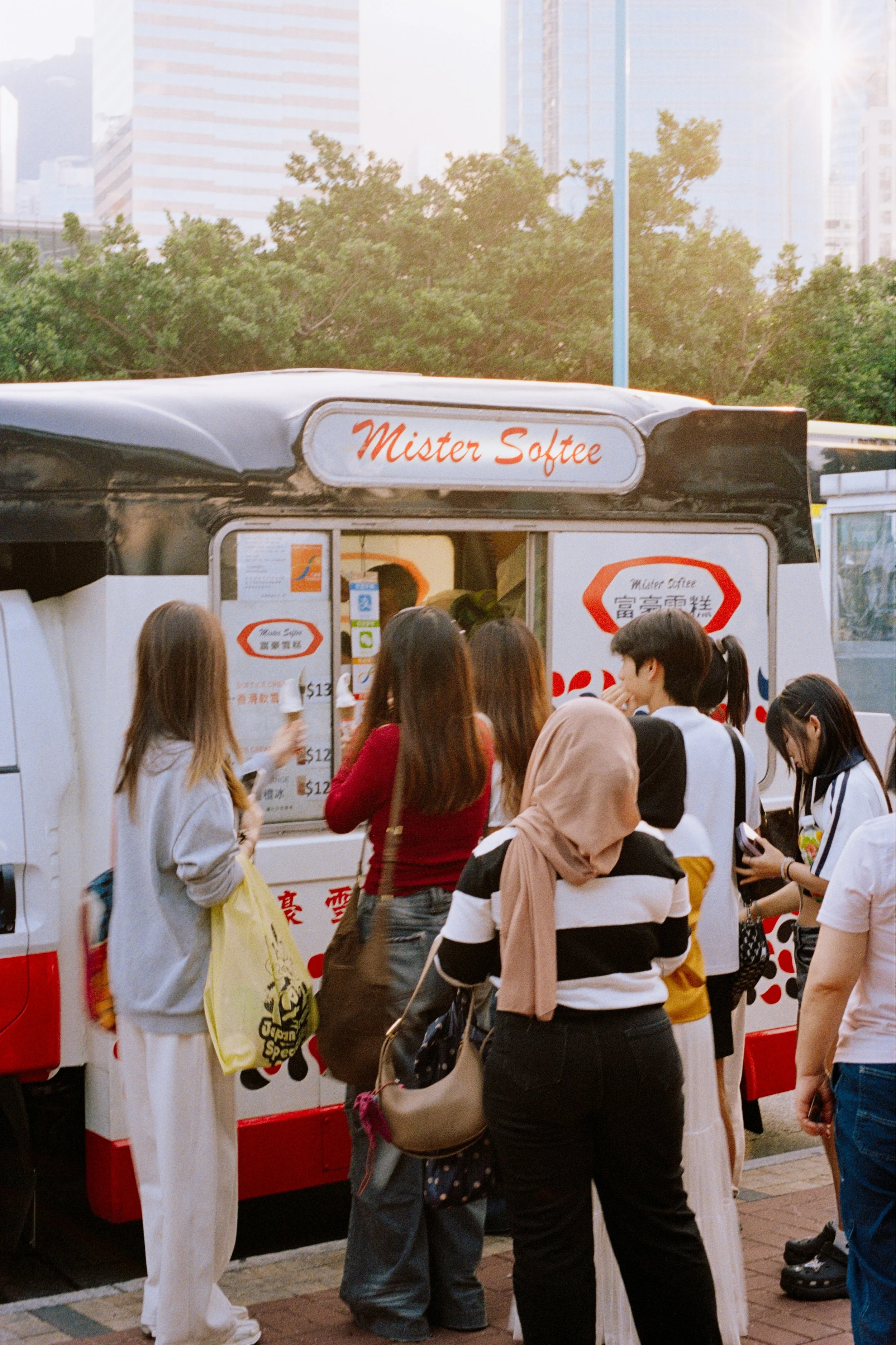 People standing in line at an ice cream truck in an urban area with trees and tall buildings in the background.