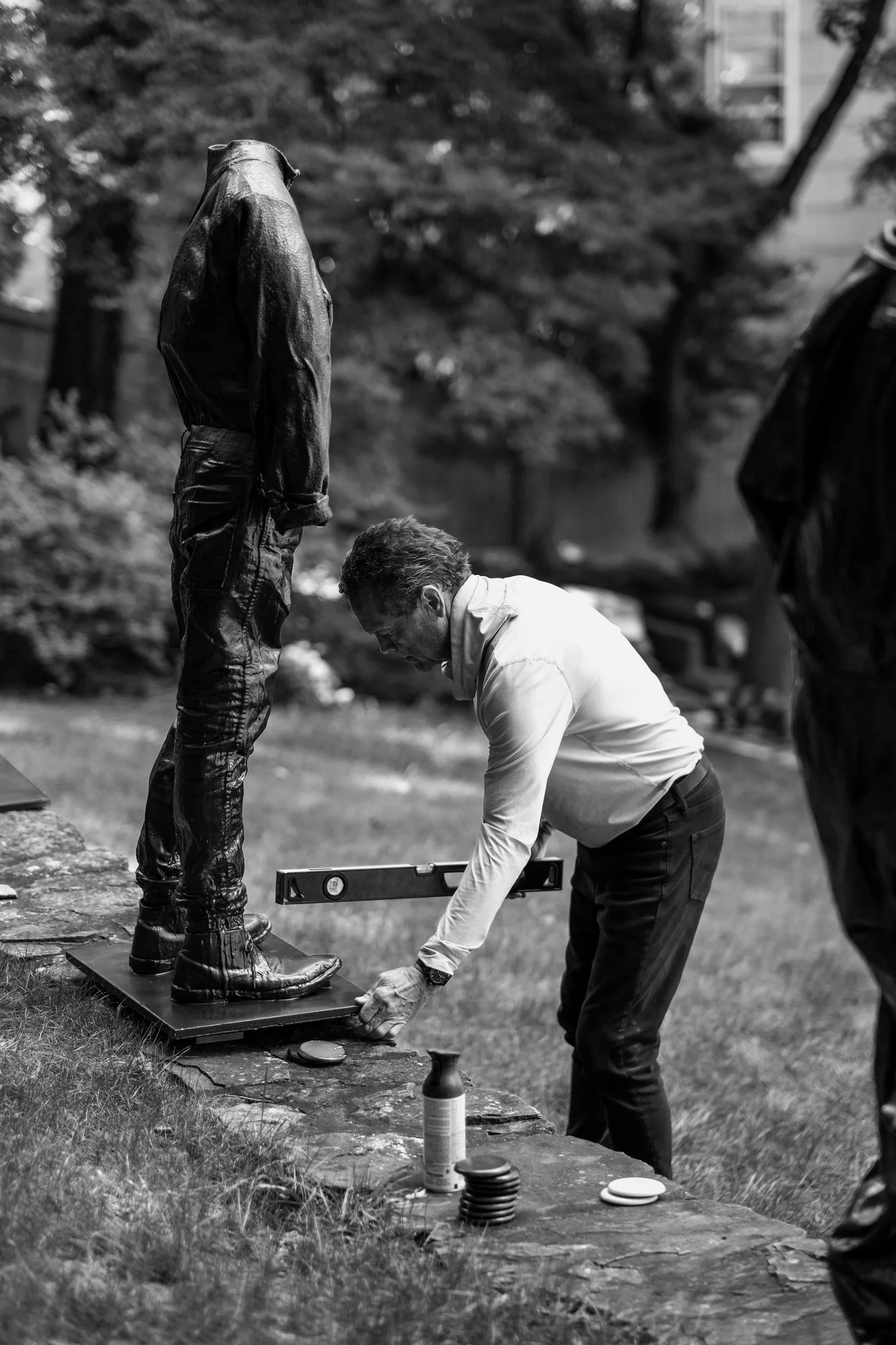 A man dressed in a white shirt and dark pants is measuring the foot of a sculpture of a person wearing boots, which is placed on a flat surface outdoors, with trees and buildings in the background.