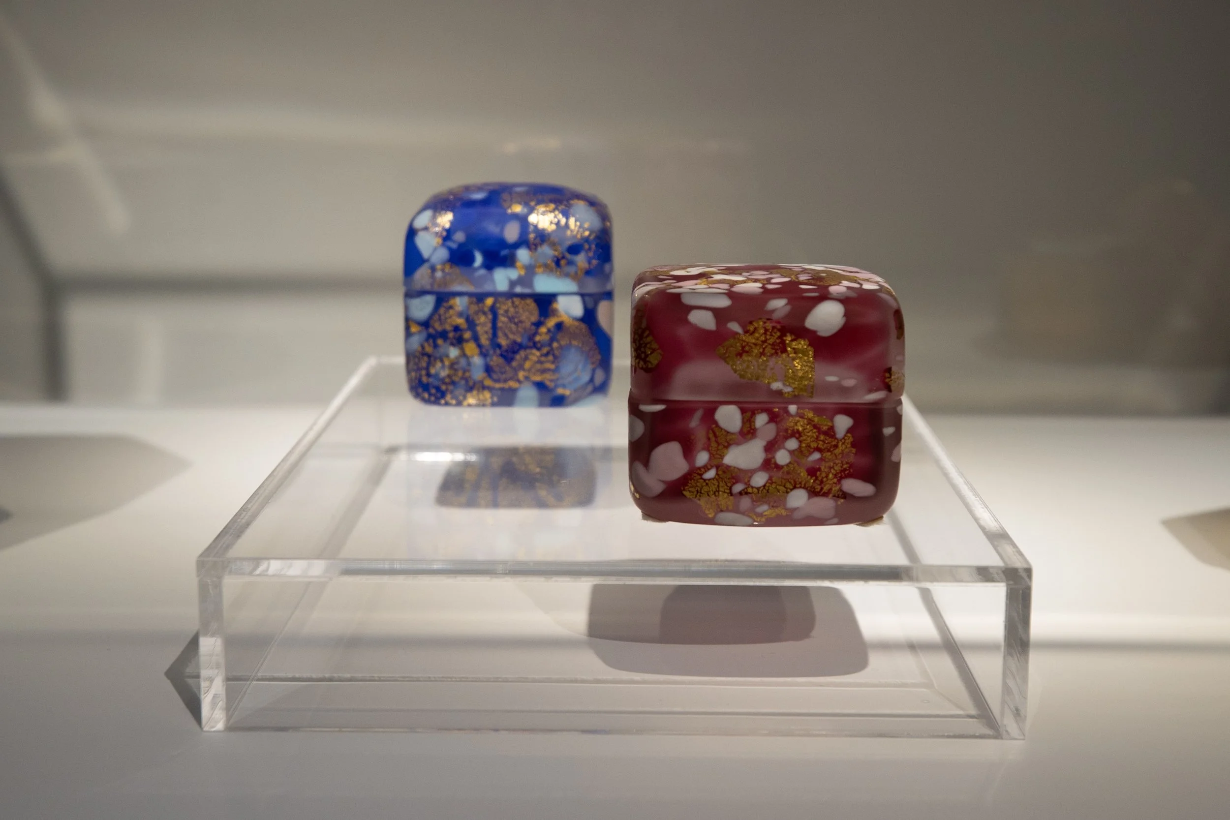 Two decorative glass cubes with gold and colorful patterns, one blue and one red, displayed on a transparent acrylic stand.