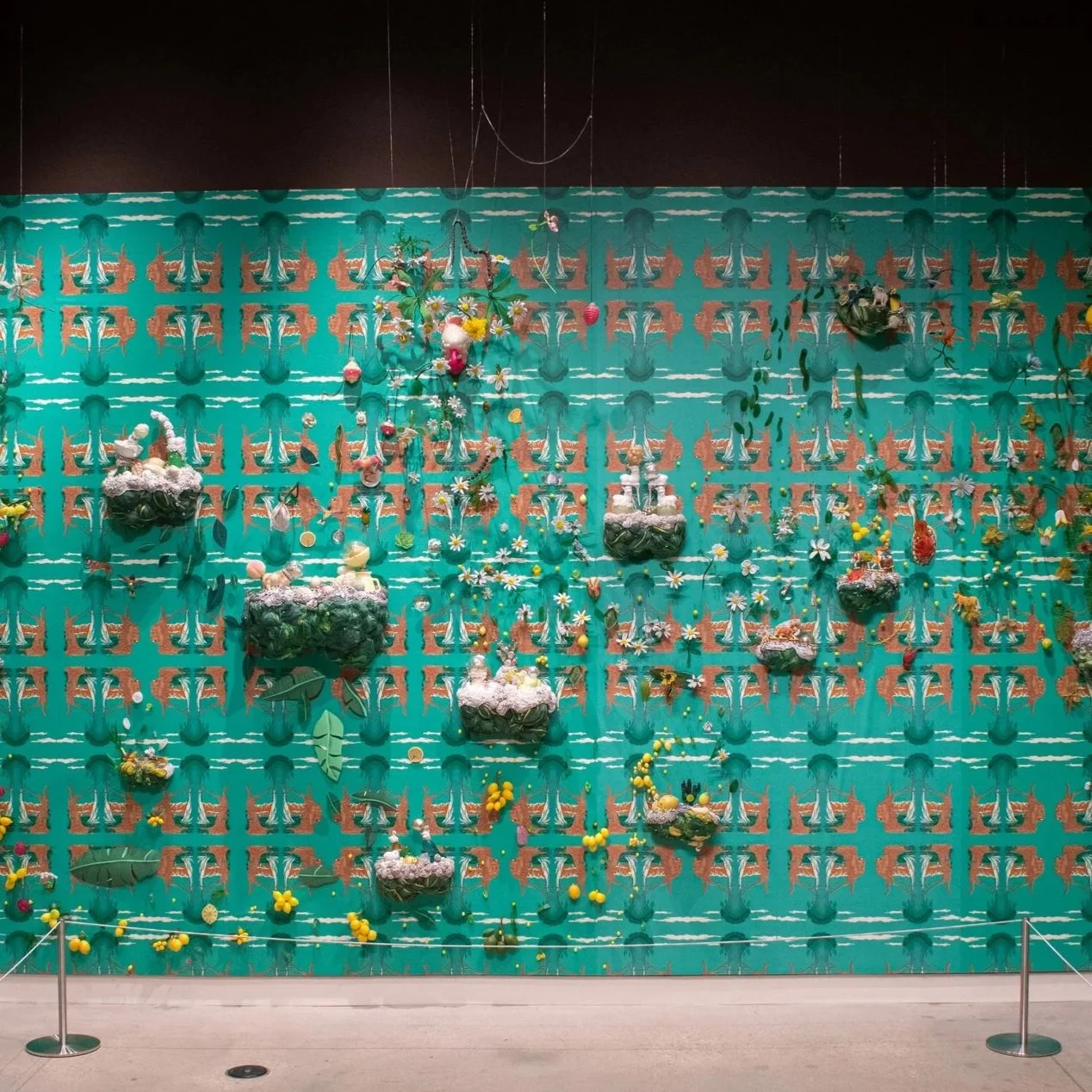 A mixed media art installation featuring found objects such as figures, set against a bright teal floral backdrop in a gallery space.