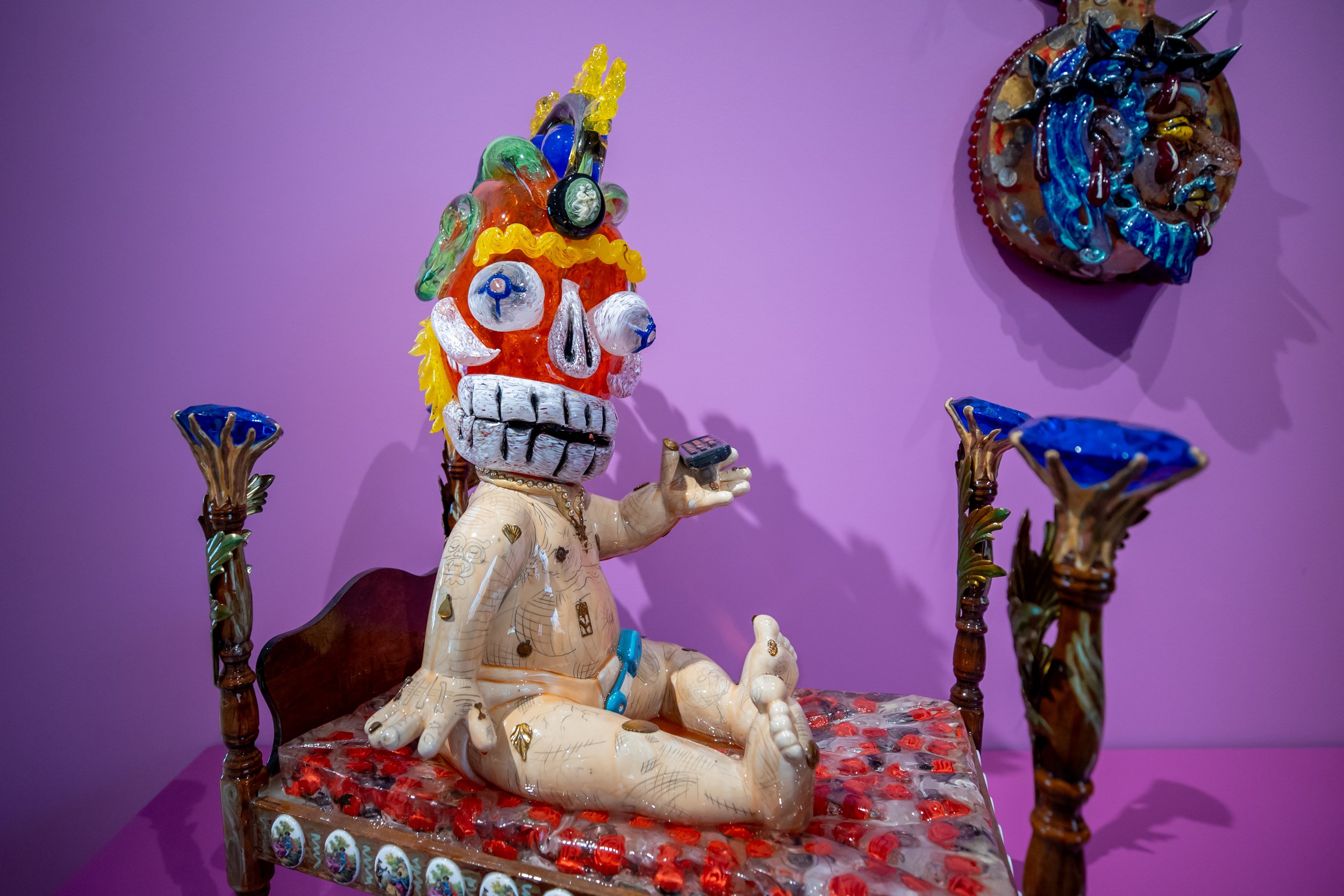 A sculpture of a doll body with a blown glass skull head against a bright purple wall.