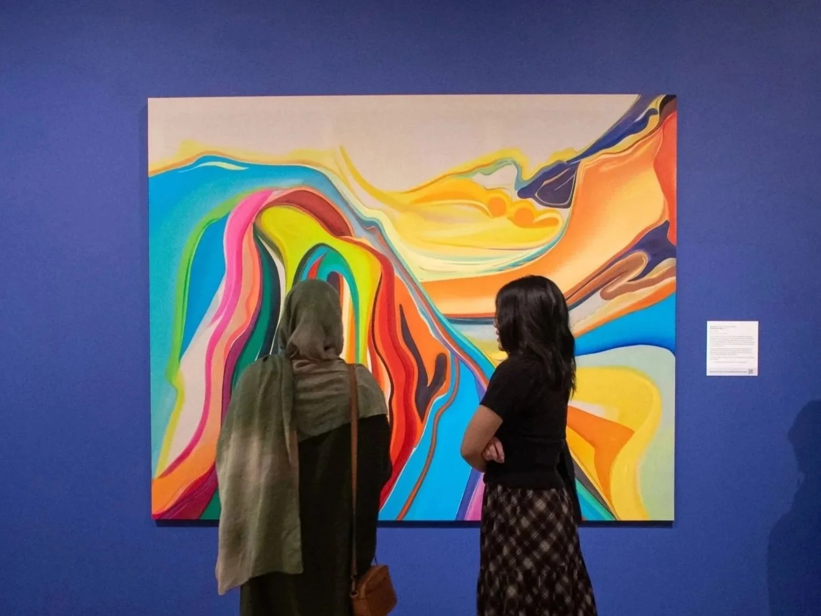 Two young people stand in front of a abstract painting hung on a blue wall.