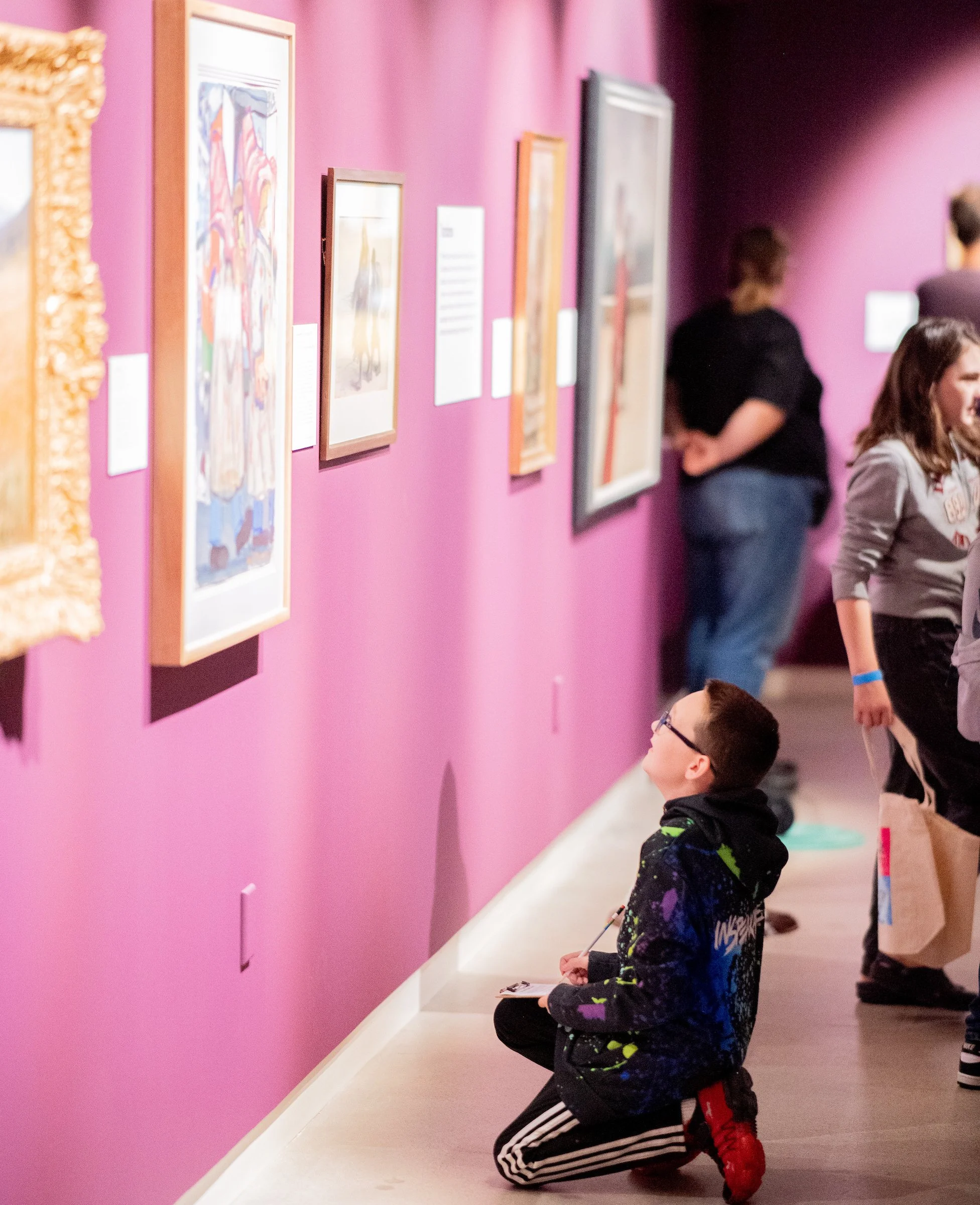 A young boy kneeling on the floor at an art gallery, looking at artwork on a pink wall, with other visitors in the background.