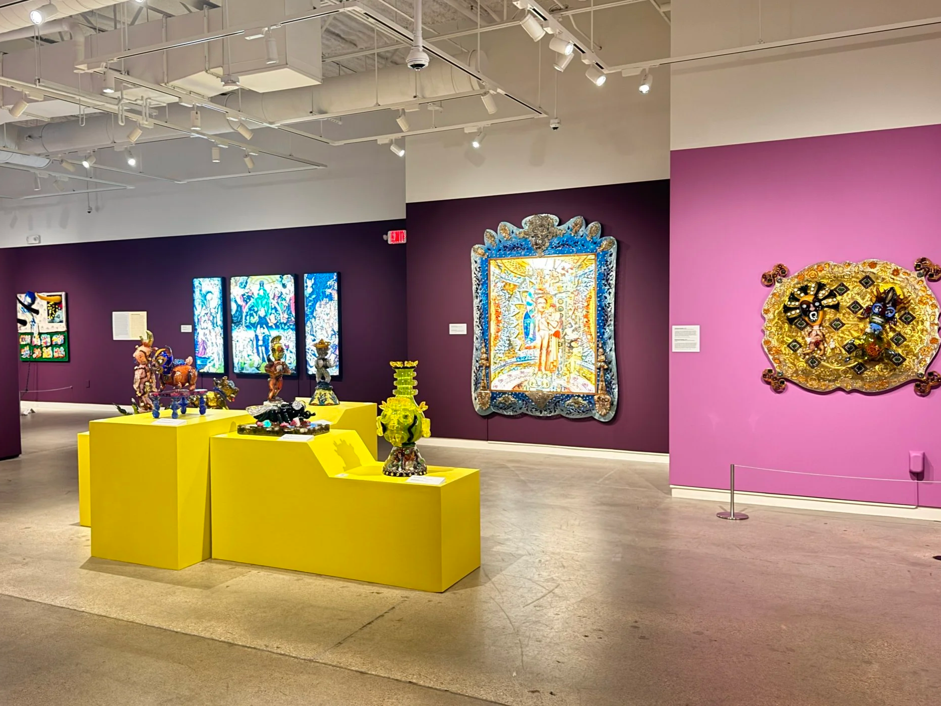 Museum or gallery exhibit displaying colorful glass artworks and decorative pieces on yellow pedestals against purple and pink walls, with spotlights overhead illuminating the displays.