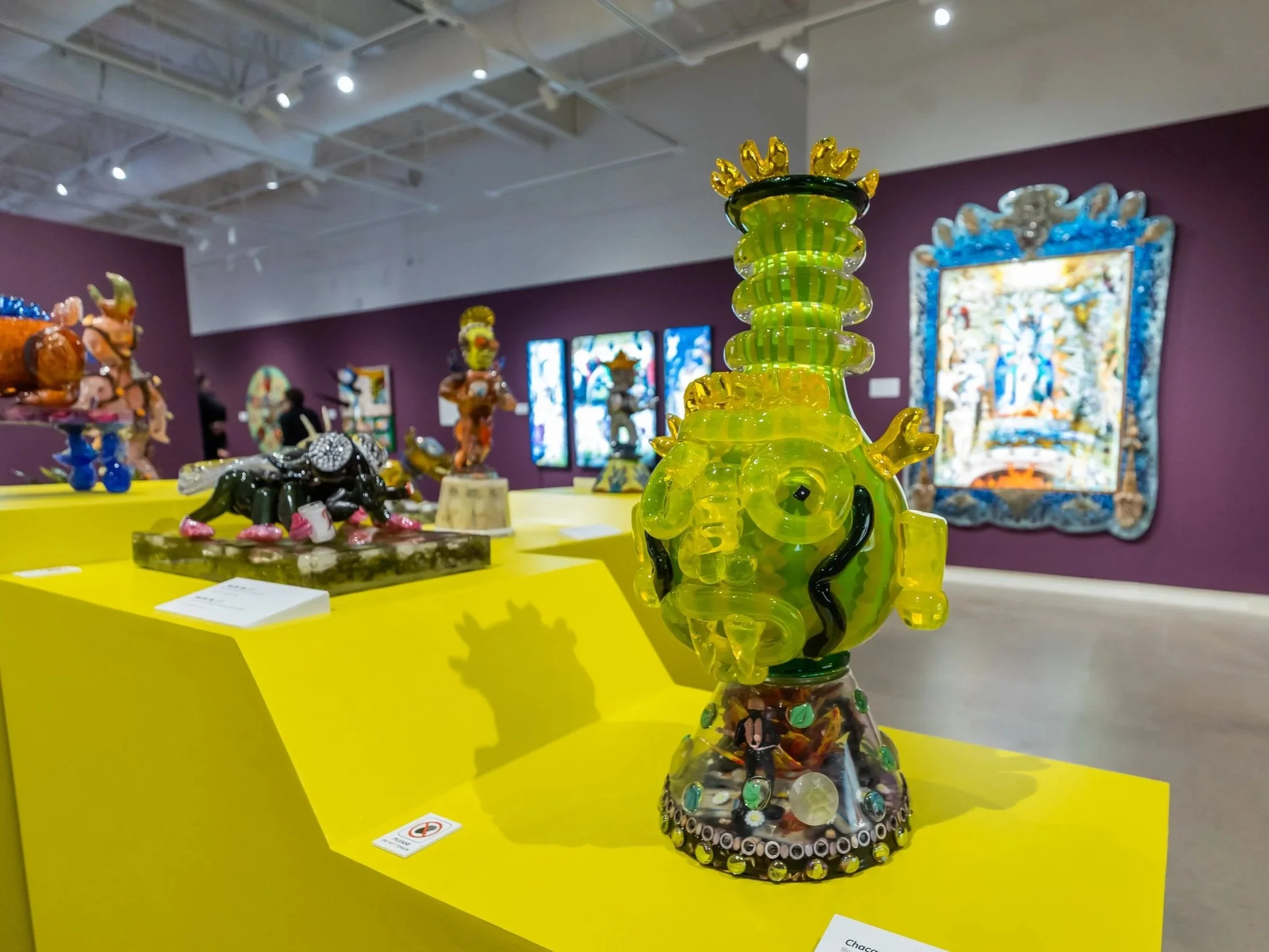 Colorful glass sculpture resembling a phallic shape, decorated with small figurines and embedded with various beads and buttons, displayed on a yellow table in an art gallery.
