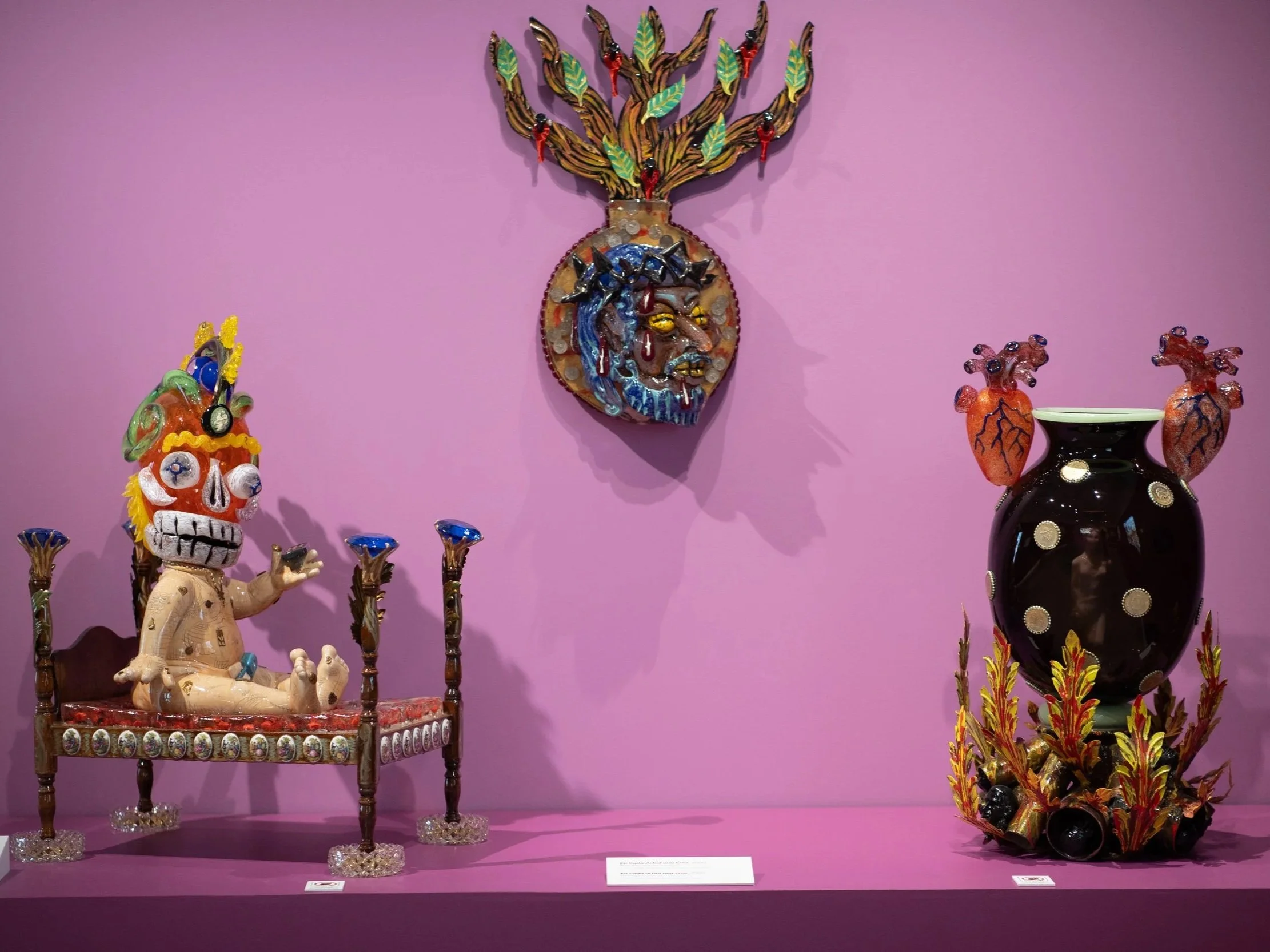 Colorful artwork in Museum of Mexican Art, pink wall background, includes a decorated skeleton doll sitting on a chair, a large mask with a face and trees or branches with green leaves on the wall, and two decorated vases with fiery designs.