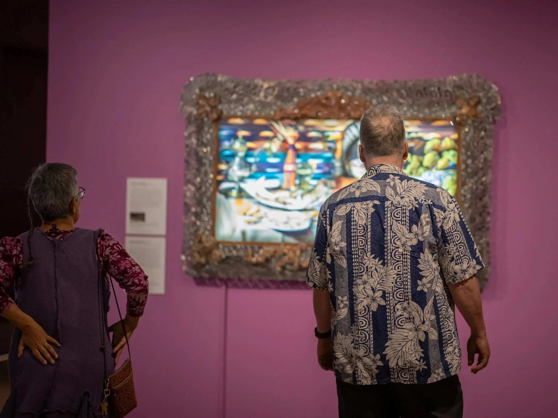 Two people are standing in front of a colorful, abstract artwork in a gallery with pink walls. One person is wearing a Hawaiian shirt and the other a purple patterned outfit with glasses.