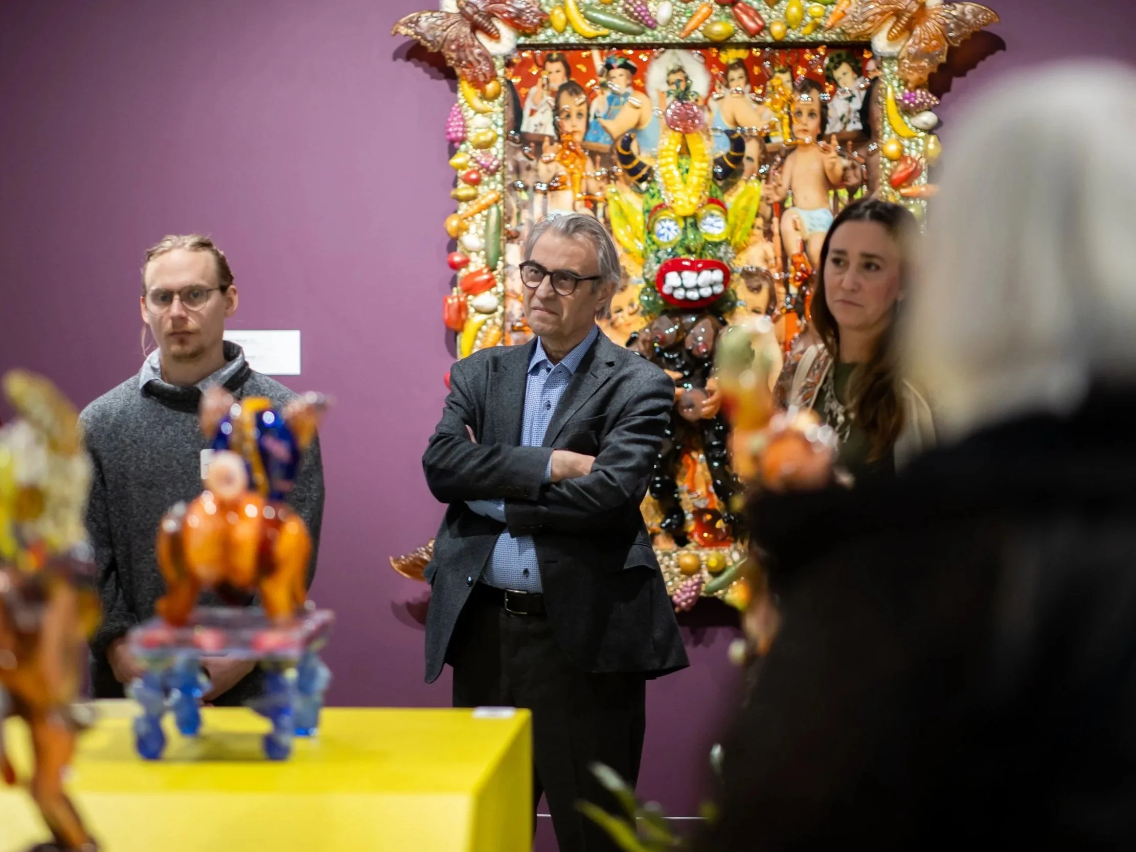 People standing in front of a colorful, decorative artwork that features a large face with exaggerated eyes and lips, surrounded by smaller figures and intricate details, in an art gallery or museum setting.