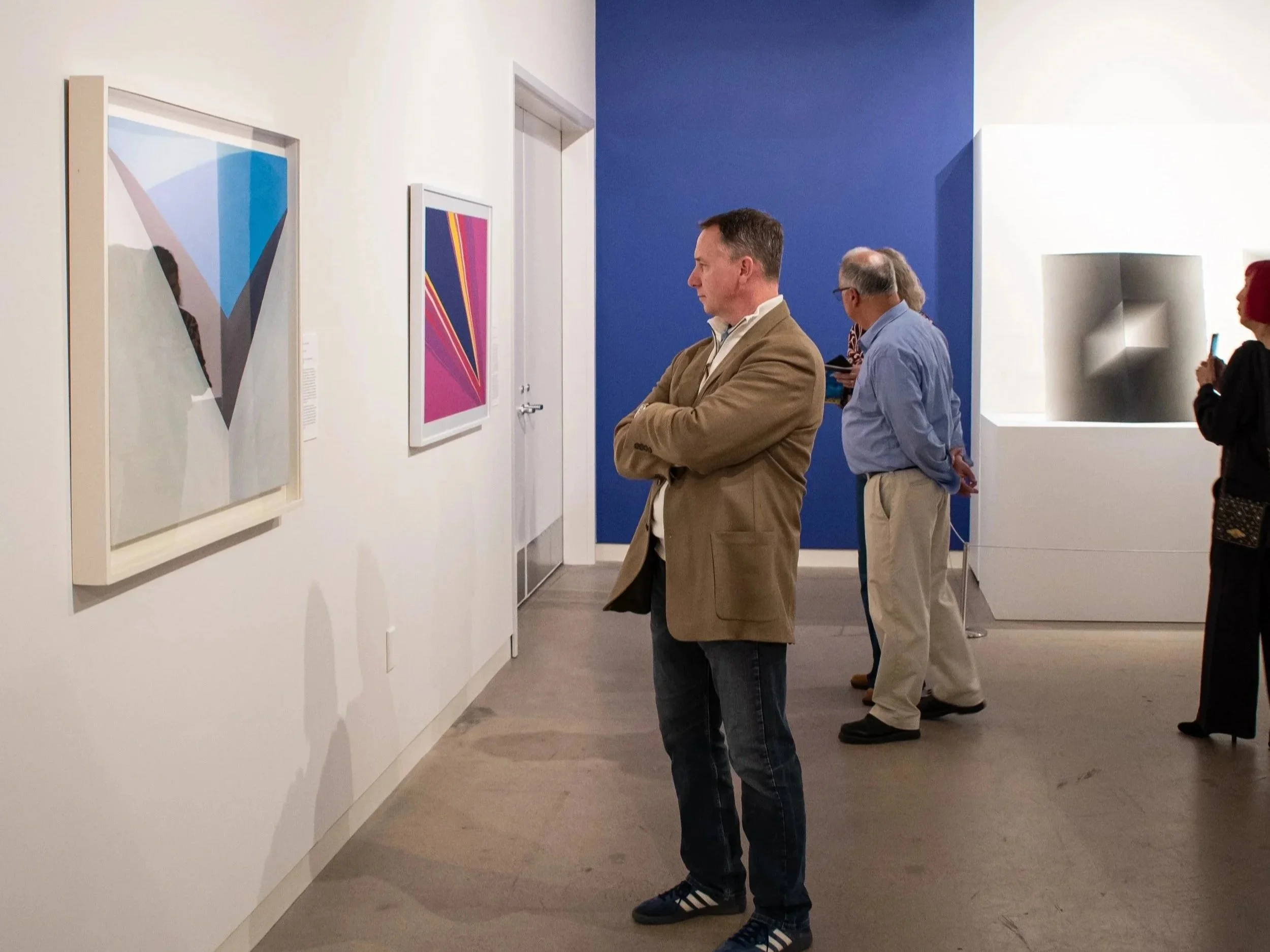 People viewing abstract artwork in an art gallery with white walls and colorful framed paintings.