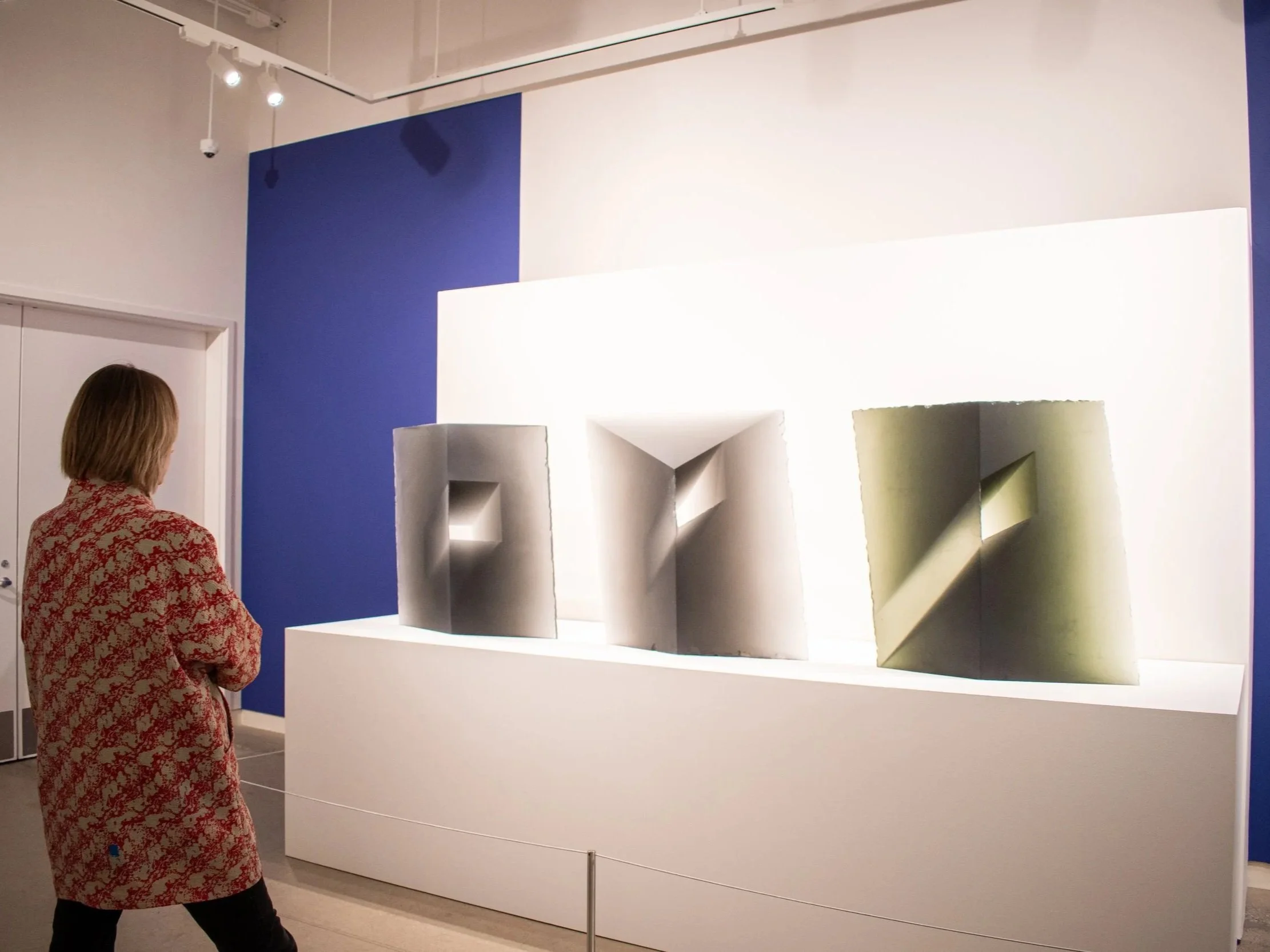 A woman wearing a patterned red and white dress is standing and observing a modern art installation in a gallery with beige walls and spot lighting.