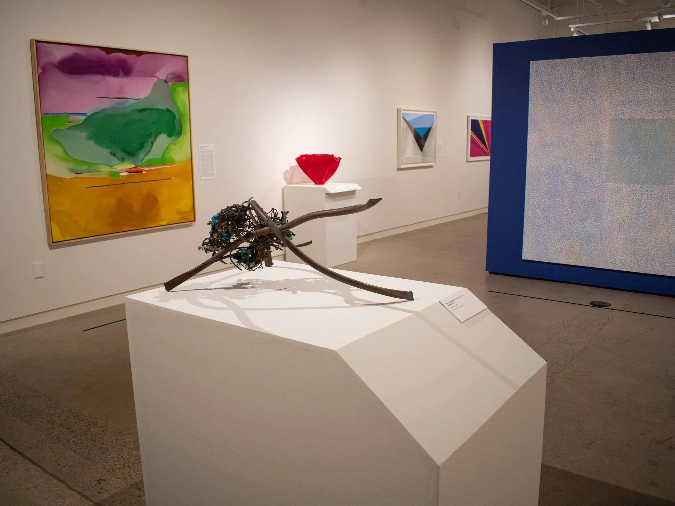 Art gallery featuring abstract paintings, a sculpture made of branches and wire, and a large mixed media piece with pixelated design.