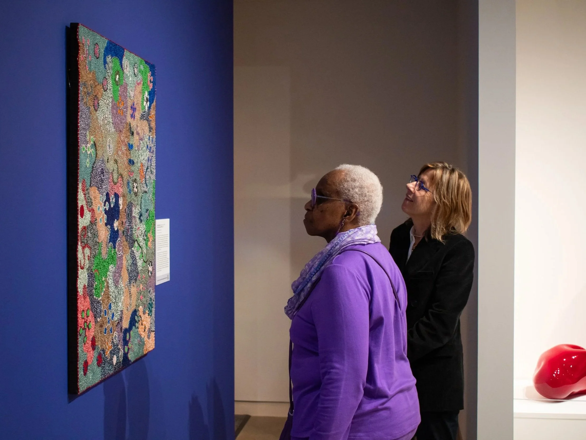 Two women observing a colorful abstract artwork on a blue wall in an art gallery.