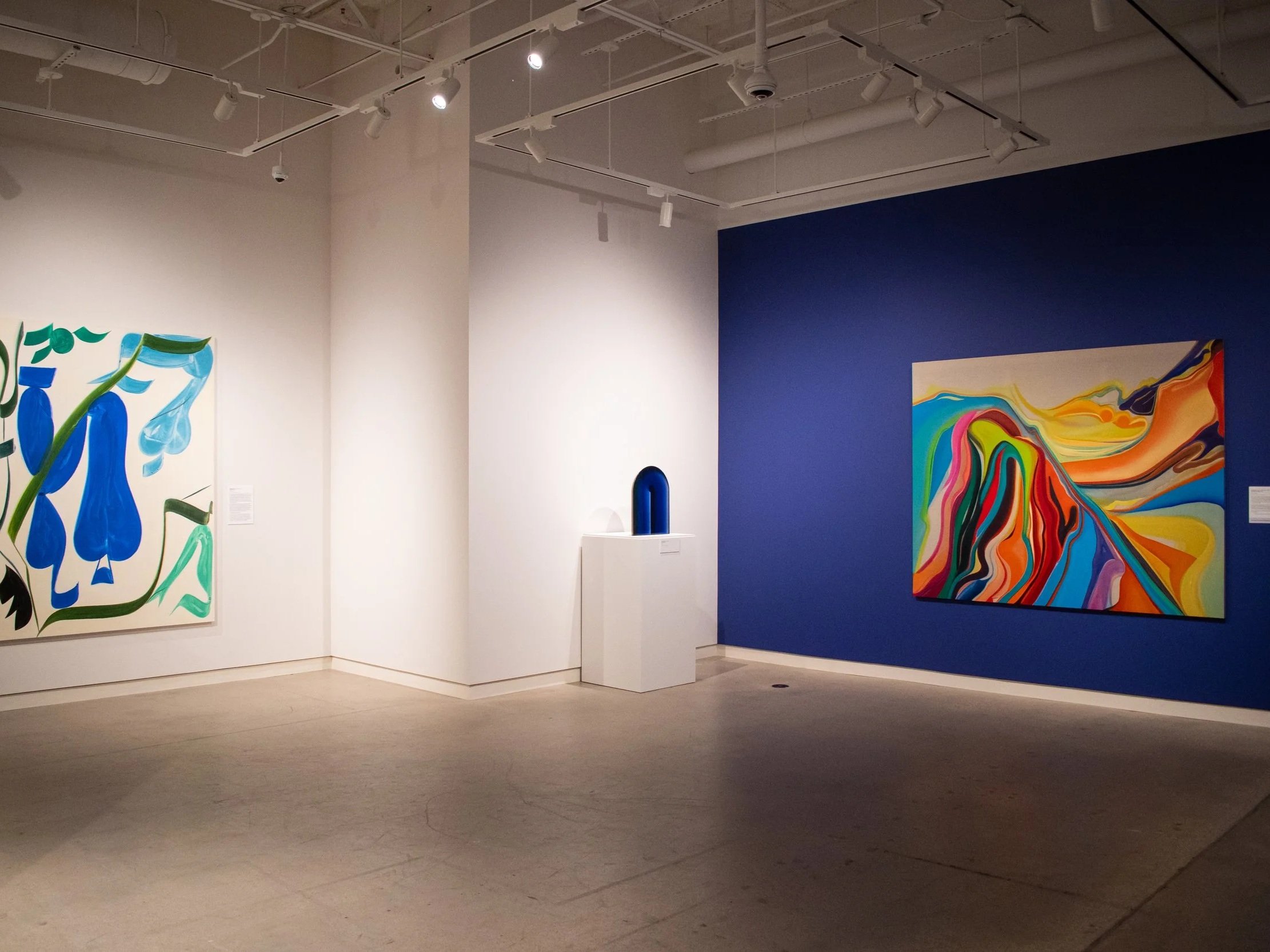 An art gallery with colorful abstract paintings on white and blue walls, ceiling track lighting, and a wooden floor.