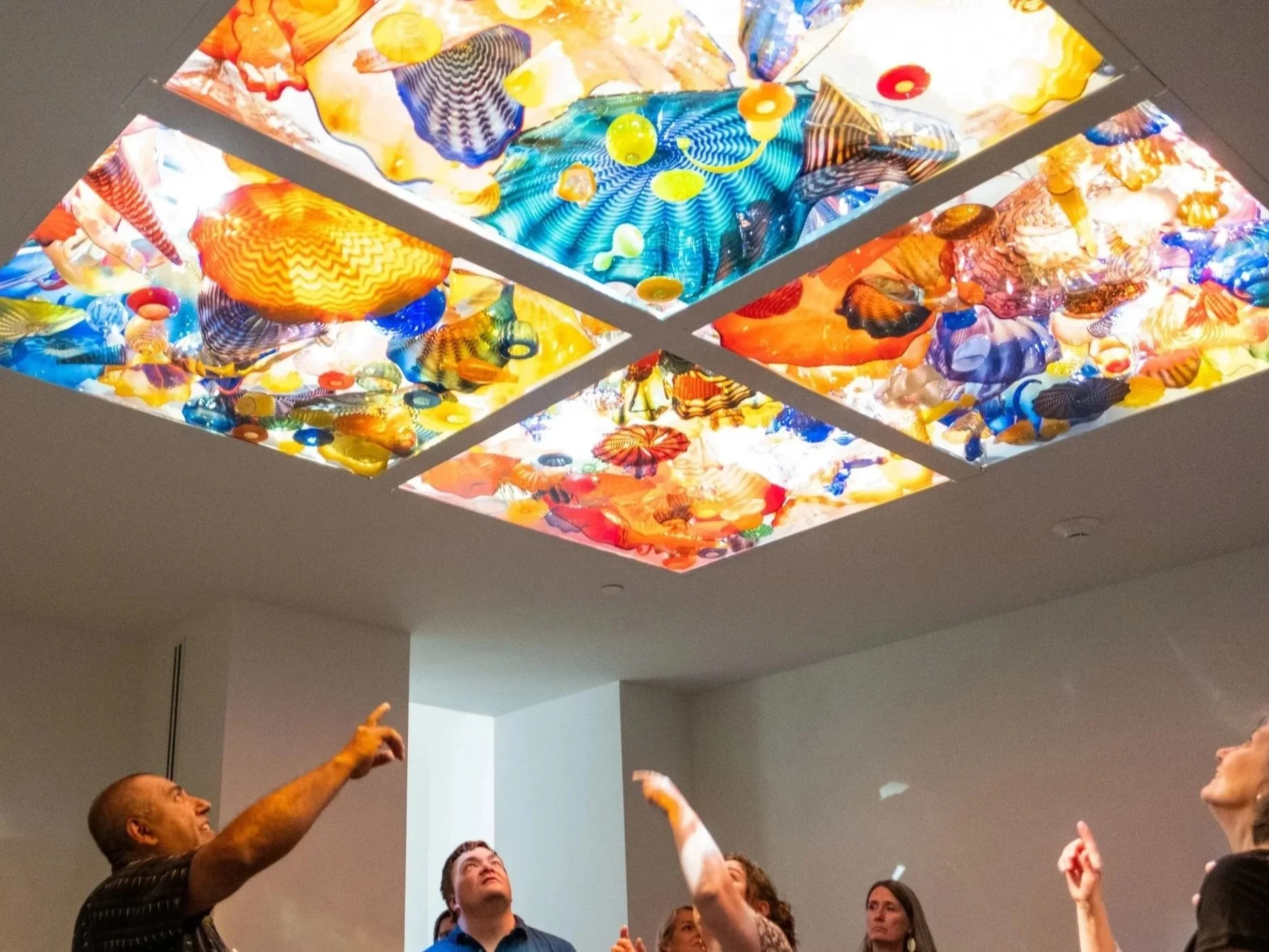 Group of people standing under a colorful, illuminated ceiling installation made of multicolored glass pieces.